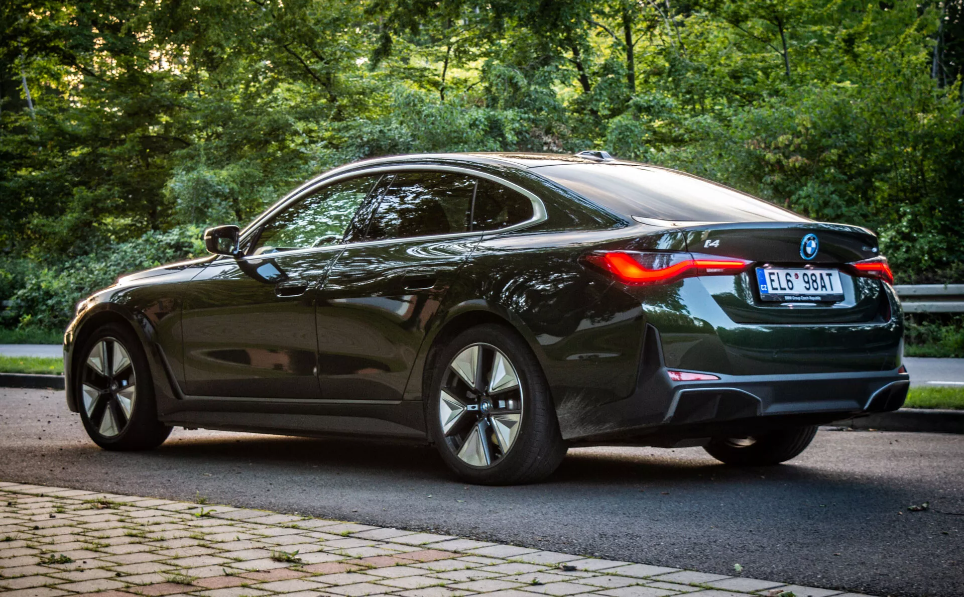 Lesklé čierne kupé BMW i4 z roku 2021 v luxusnom zobrazení z troch štvrtín zozadu, predvádzajúce športové viacramenné zliatinové kolesá, LED zadné svetlá a panoramatickú strechu uprostred zelenej krajiny obklopenej stromami vo dne