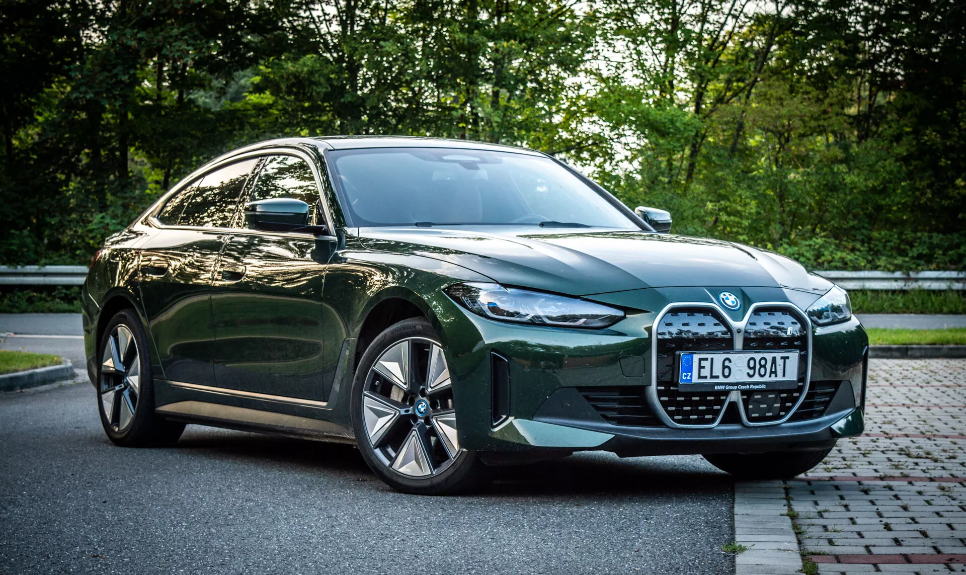 Zelený štvordverový sedan BMW i4 z roku 2021 zaparkovaný na tehlovej ulici vedľa trávnatej plochy s listím na pozadí, so záberom z predného štvrťuha ukazujúcim veľkú mriežku auta, LED svetlomety, vzduchové vstupy a zliatinové kolesá