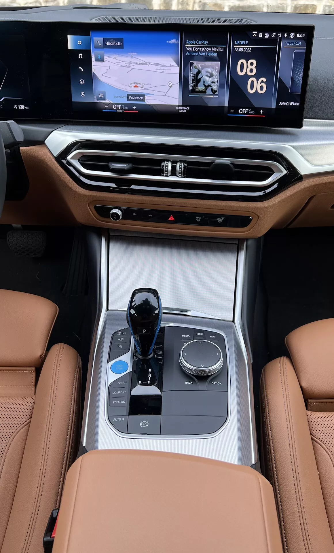 Pohľad na stredovú konzolu interiéru BMW i4 z roku 2021 zo zadných sedadiel, zdôrazňujúci kožené sedadlá, elektronický radenie prevodovky, ovládací gombík multimédií, držiaky nápojov, ovládanie klímy a dvojrozmerný infotainment displej s navigáciou a zobrazením médií