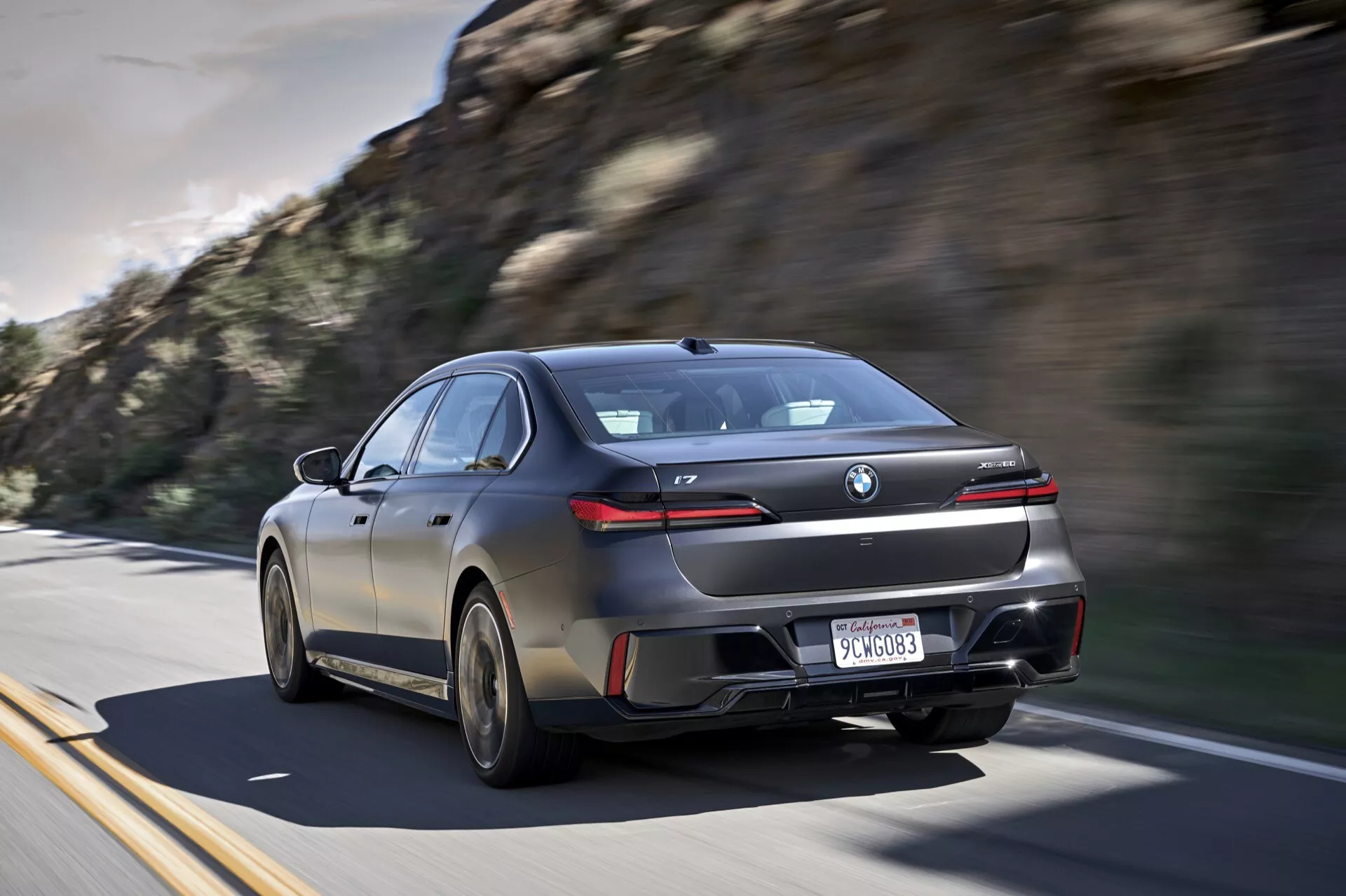 Moderný sedan BMW i7 (G70) z roku 2022 v pohybe na diaľnici, ukazujúci jeho športový kupé-podobný siluetu, výrazné LED zadné svetlá cez celý kufor, veľké zliatinové kolesá a výfukový systém, v tmavom lesklom prevedení s tónovanými oknami