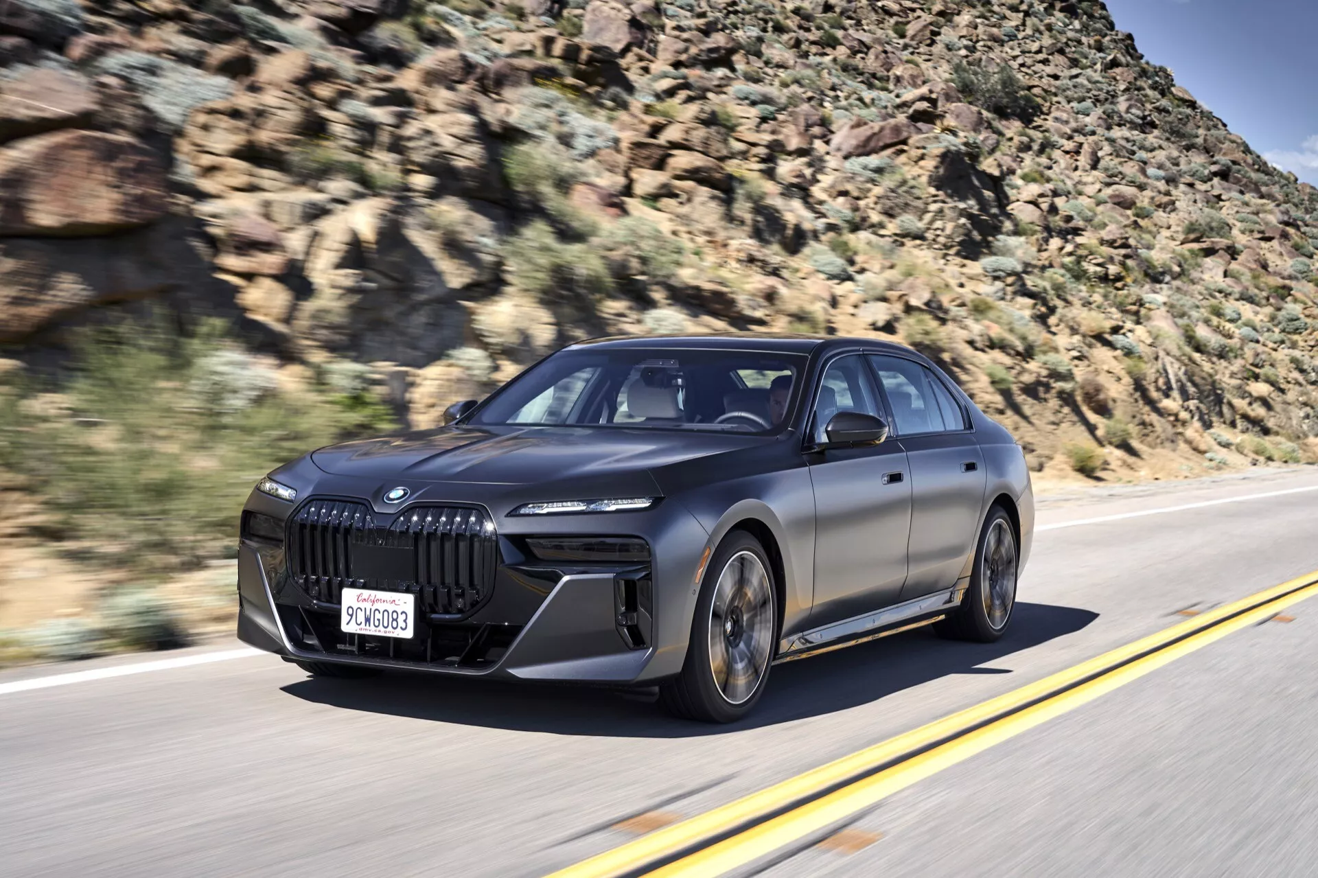 Dynamický záber z predného trojštvrtečného uhla BMW i7 (G70) z roku 2022 v pohybe, zdôrazňujúci jeho veľkú obličkovitú mriežku, LED svetlomety a elegantné dizajnové prvky na pozadí rozmazanej cesty a skalnatej krajiny.
