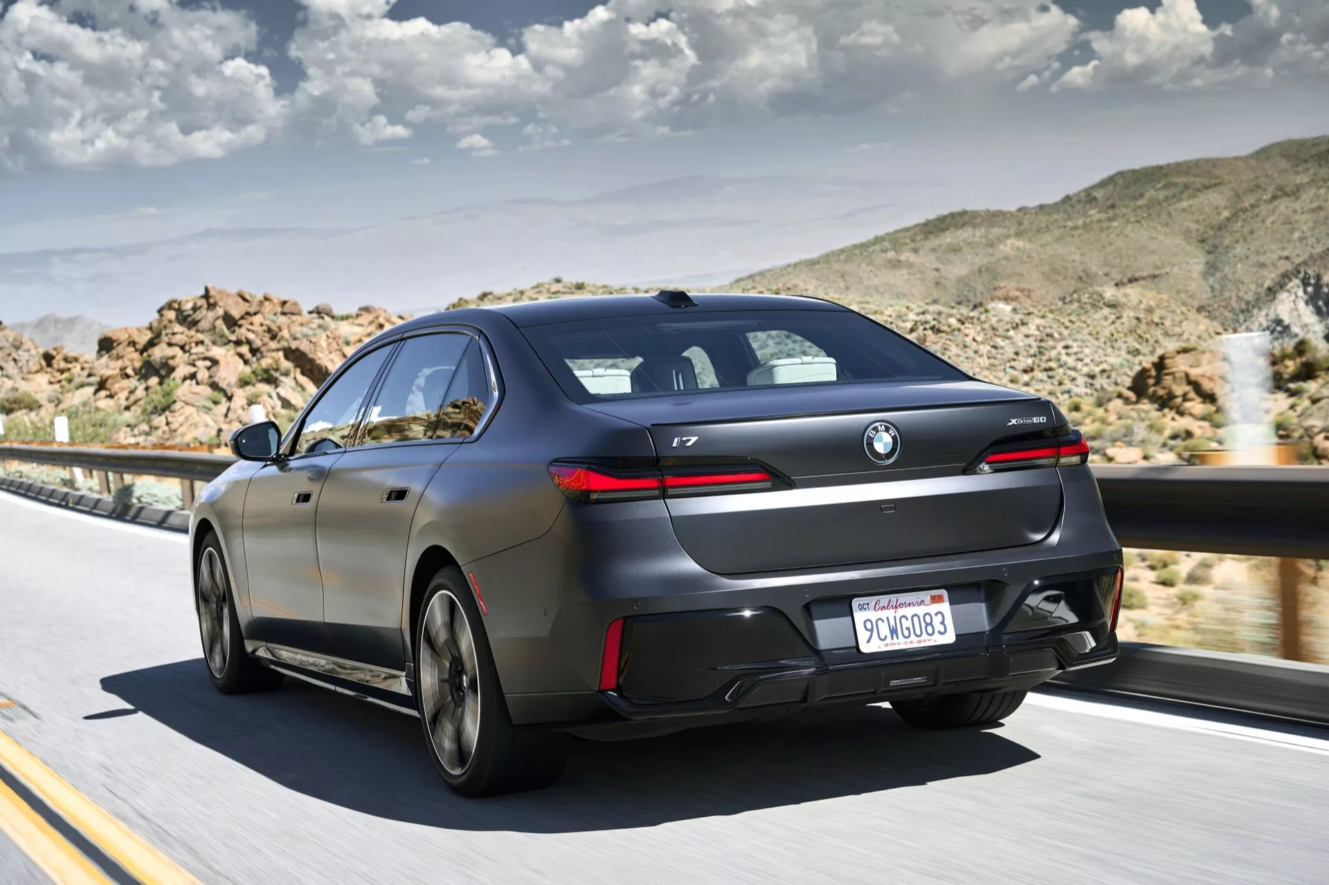 BMW i7 (G70) 2022 v pohybe, zadný trojštvrťový pohľad zvýrazňujúci zadné svetlá, zadný nárazník a aerodynamické bočné línie proti púštnej krajine