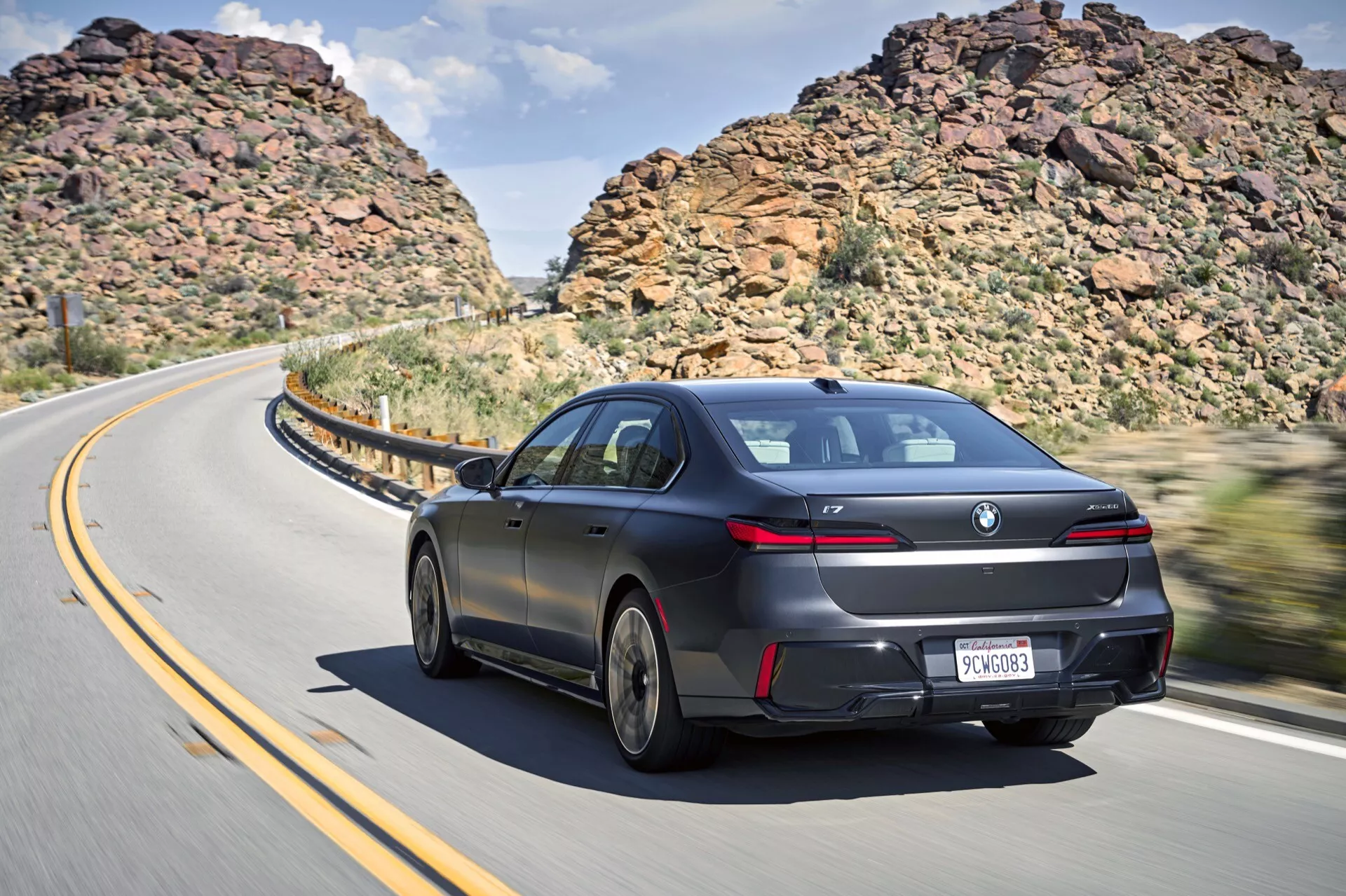 Dynamický zadný trojštvrťový pohľad na sedan BMW i7 2022 (G70) v pohybe na zákrutej ceste so skalnatými svahmi a čiastočne zamračeným nebom, predvádzajúci elegantný aerodynamický dizajn, lesklý lak, dizajnové zliatinové kolesá, LED zadné svetlá, jemný zadný spojler, tónované okná a dvojité výfukové otvory.