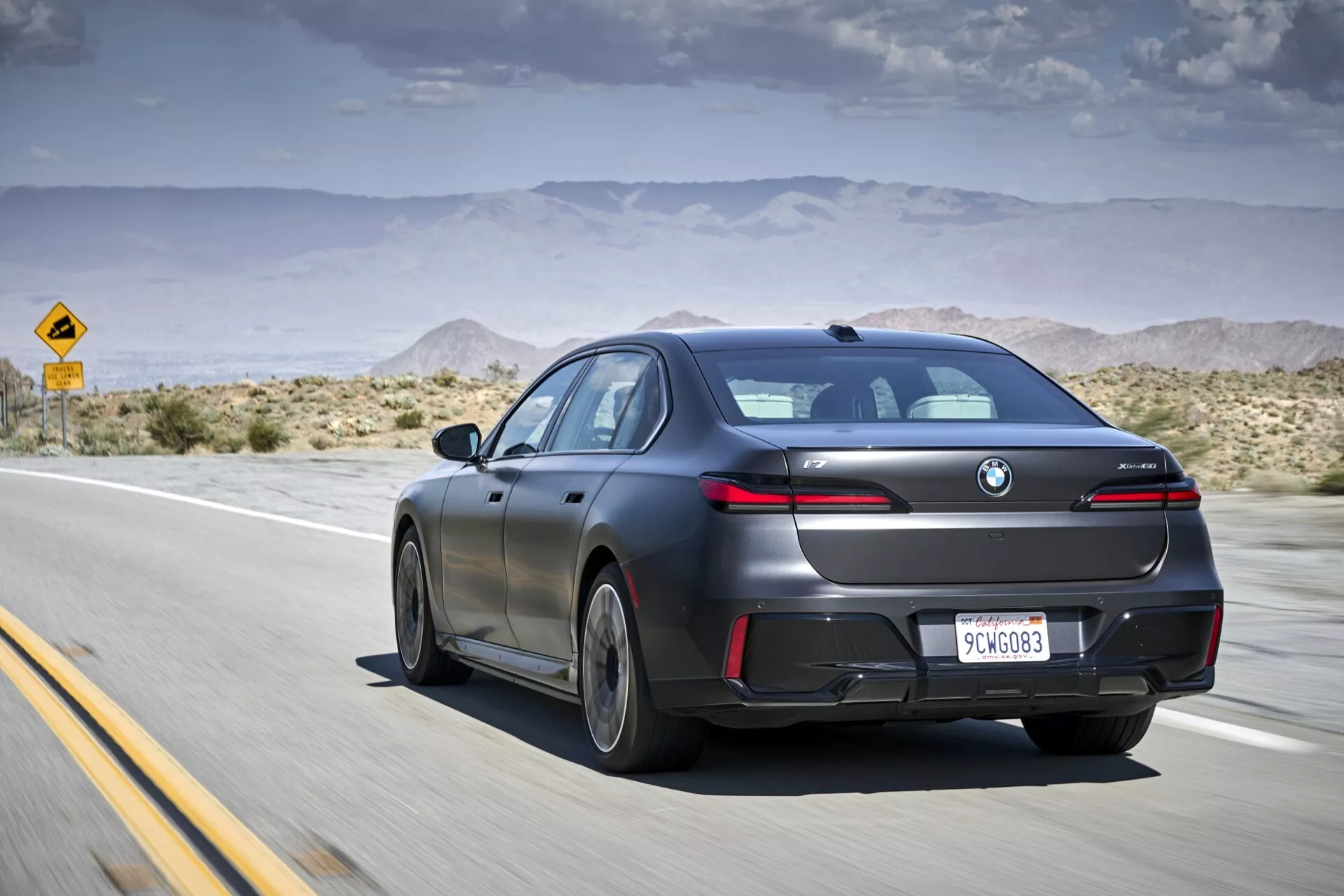 Moderný sedan BMW i7 (G70) z roku 2022 v matnom prevedení zachytený v pohybe z vonkajšieho, zadného trojštvrťového uhla na púštnej diaľnici, ukazujúci jeho aerodynamický dizajn, LED zadné svetlá a technologické funkcie, ako sú parkovacie senzory a strešné okno, pod čiastočne zamračenou oblohou.
