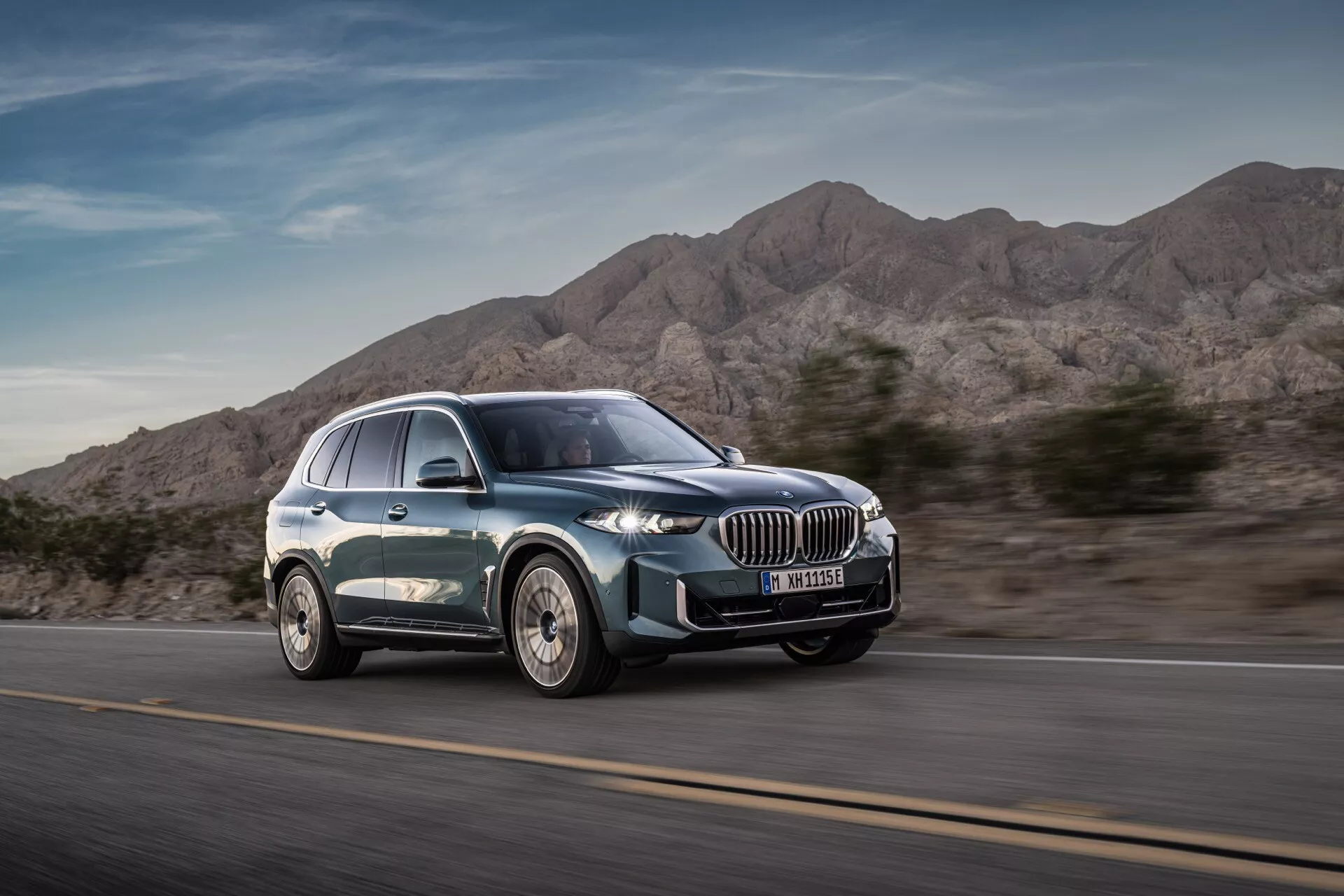 Moderné luxusné SUV BMW X5 2023 (G05-F95 Facelift) v pohybe z uhla pohľadu tri štvrtiny spredu zdôrazňujúce dynamický dizajn, veľkú obličkovitú mriežku, úzke LED svetlomety, zliatinové kolesá s viacramenným dizajnom a hladký siluetu s metalickým lakom