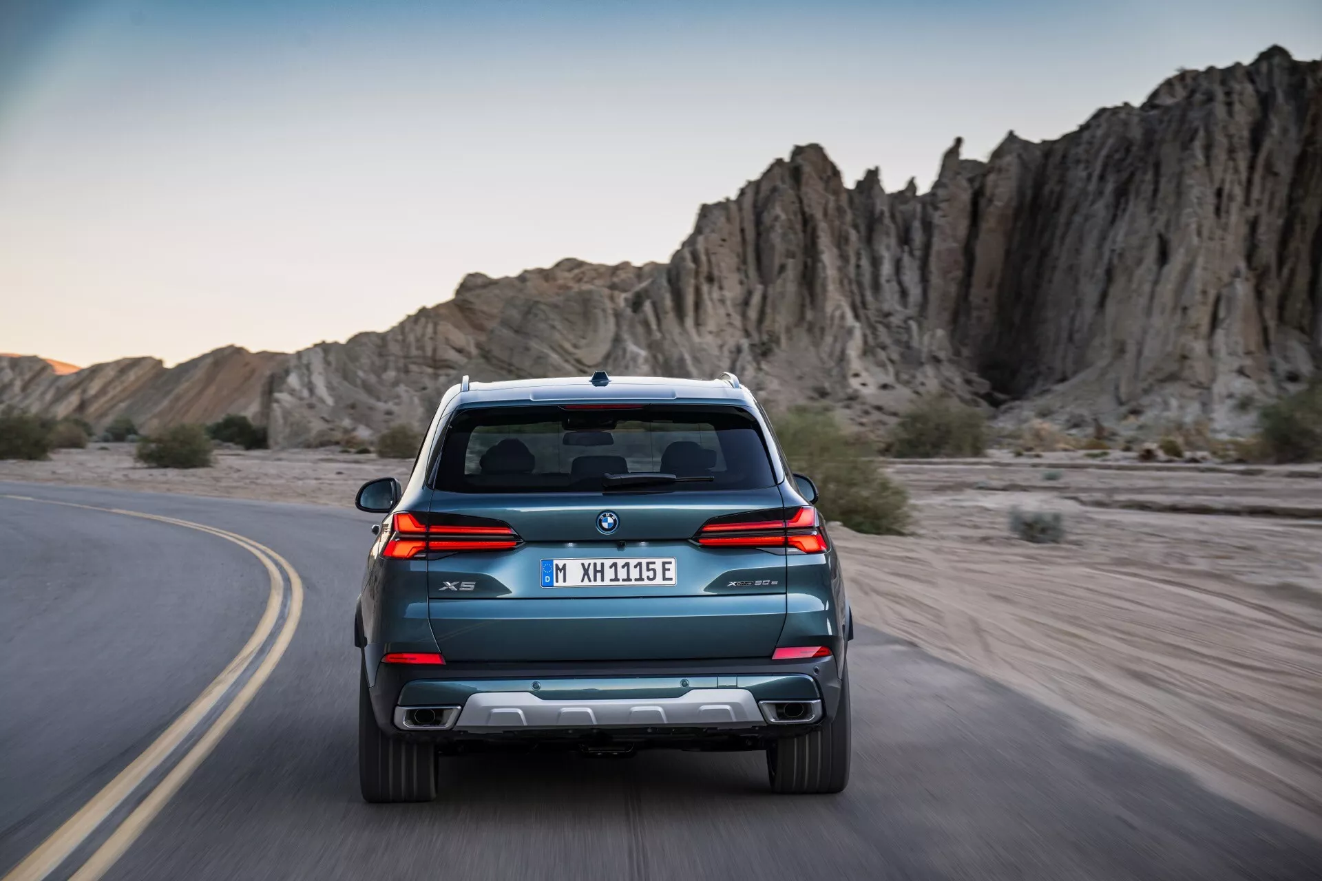 Zadný pohľad na BMW X5 G05 F95 facelift 2023, idúce po zákrute, s elegantnými líniemi, dvojitým výfukom a parkovacími senzormi na pozadí dobrodružného skalnatého terénu
