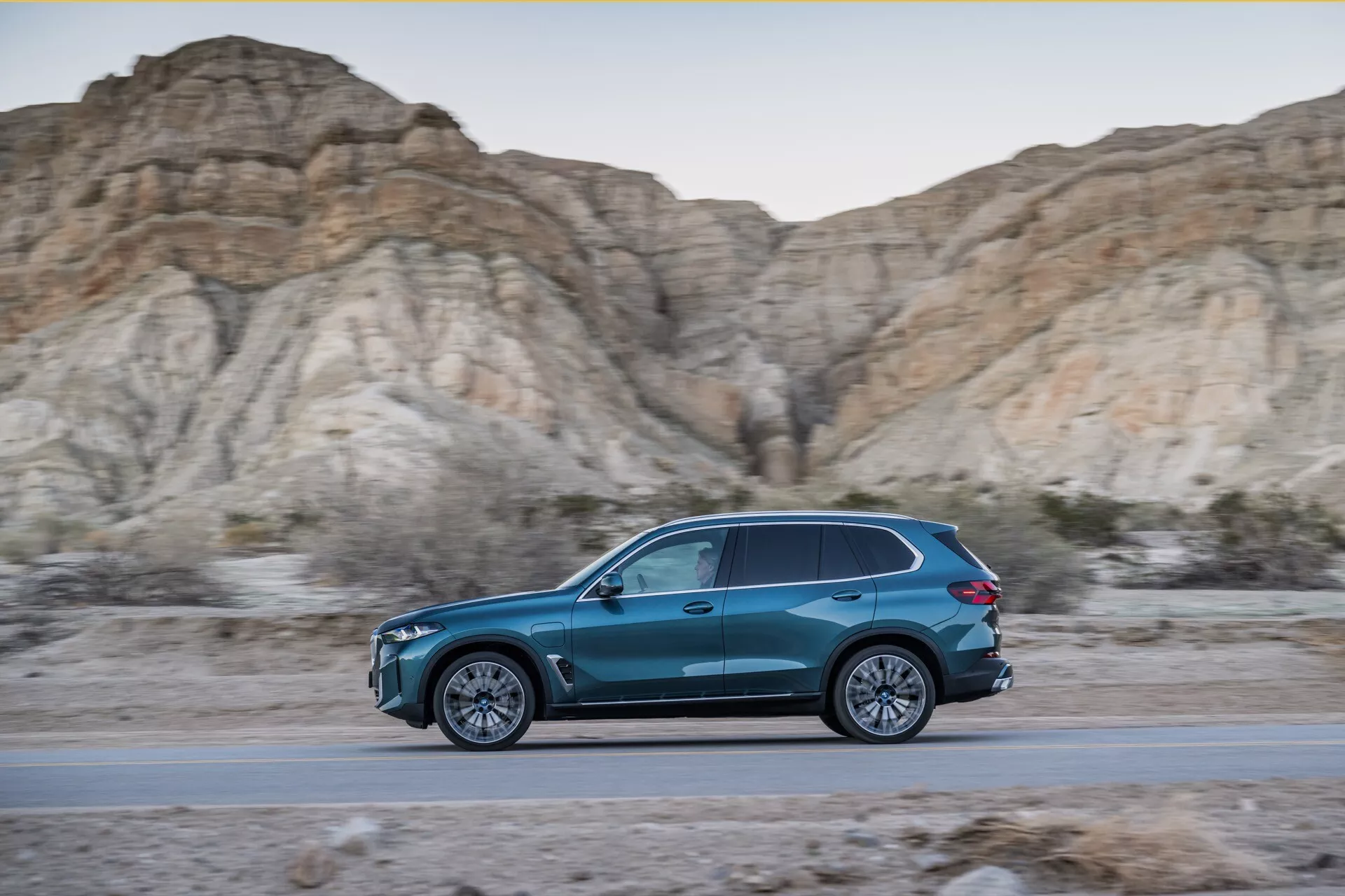 Dynamický bočný profil BMW X5 2023 (G05-F95 facelift) v pohybe, modré SUV tvaru slzy na ceste s pozadím drsných, holých kopcov počas zlatej hodiny, ukazujúci štíhly dizajn, aerodynamickú účinnosť, viacdielne zliatinové kolesá a LED osvetlenie