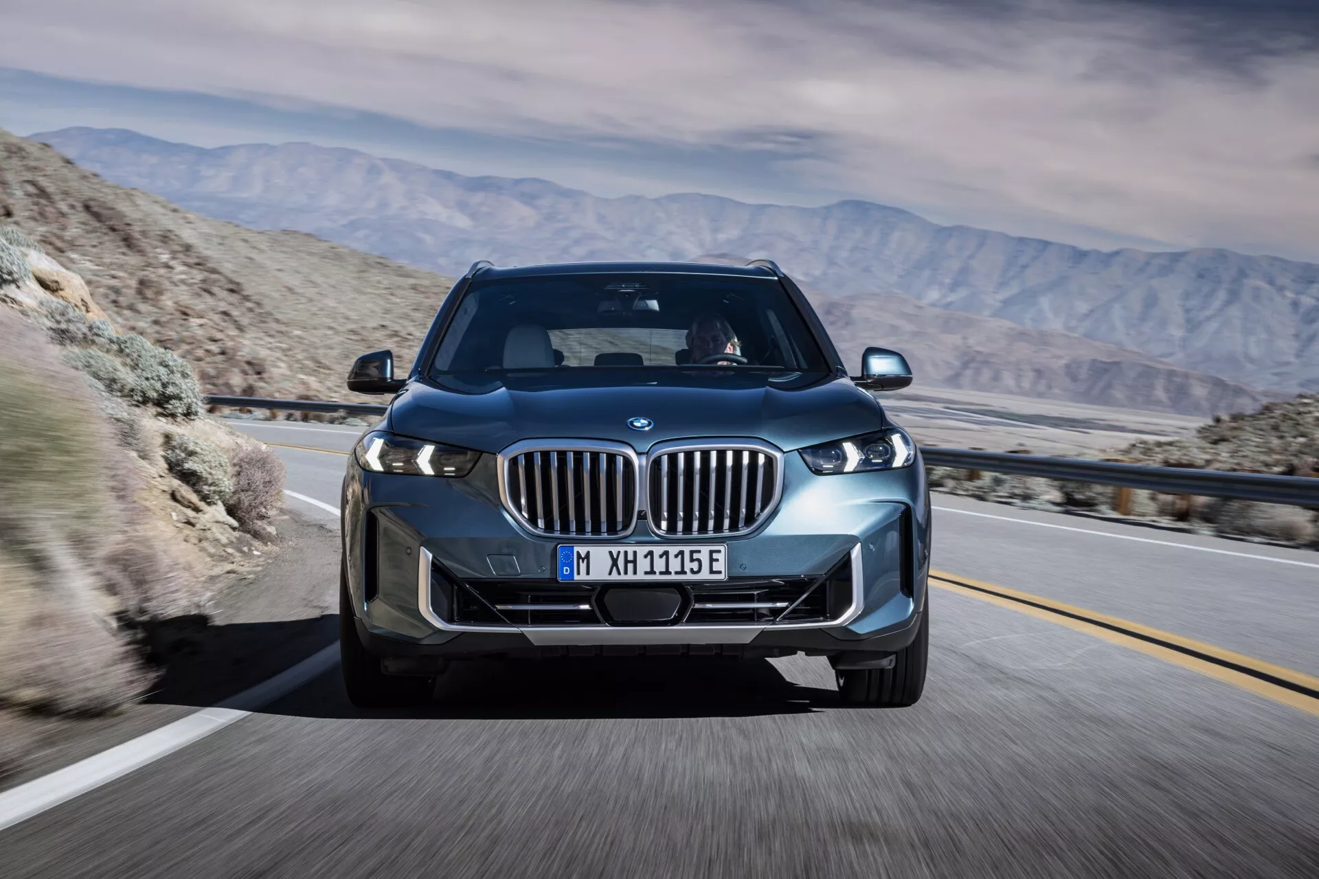 BMW X5 2023 (G05 F95 facelift) v pohybe, zachytené z predného trojštvrtečného uhla, zvýrazňujúce jeho LED svetlomety, obličkovú mriežku s aktívnymi žalúziami, zliatinové kolesá a elegantný aerodynamický dizajn proti pozadiu horskej púšte, naznačujúce pokročilú technológiu a potenciálnu elektrickú alebo hybridnú schopnosť