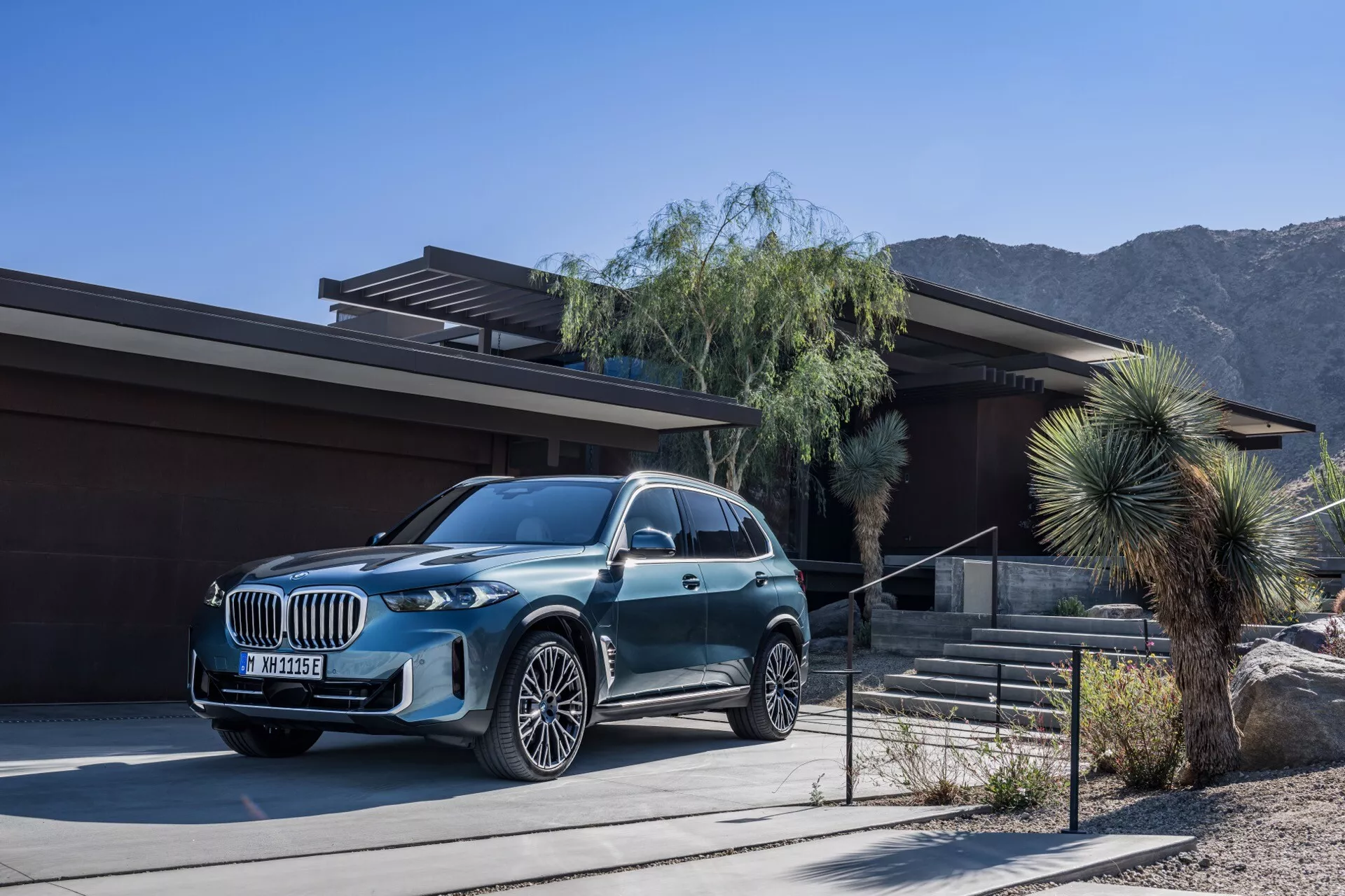 Luxusné SUV BMW X5 2023 (G05-F95 Facelift) zaparkované pred súčasným domom s dezertnou a horskou krajinou na pozadí, s kovovým povrchom, veľkou mriežkou, LED svetlometmi a viacramennými zliatinovými kolesami