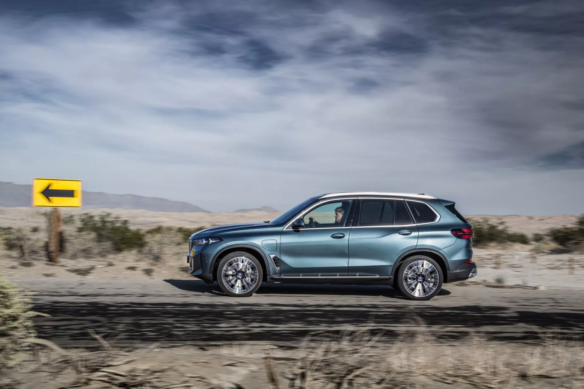 Bočný profil BMW X5 2023 (G05-F95 Facelift) v pohybe, ukazujúci jeho metalický lak, aerodynamický dizajn, luxusné zliatinové kolesá, tónované okná so zrkadlami smerových svetiel, strešné lyžiny a pokročilú LED technológiu, na pozadí púšte s dynamickou žltou značkou zatáčky vľavo.