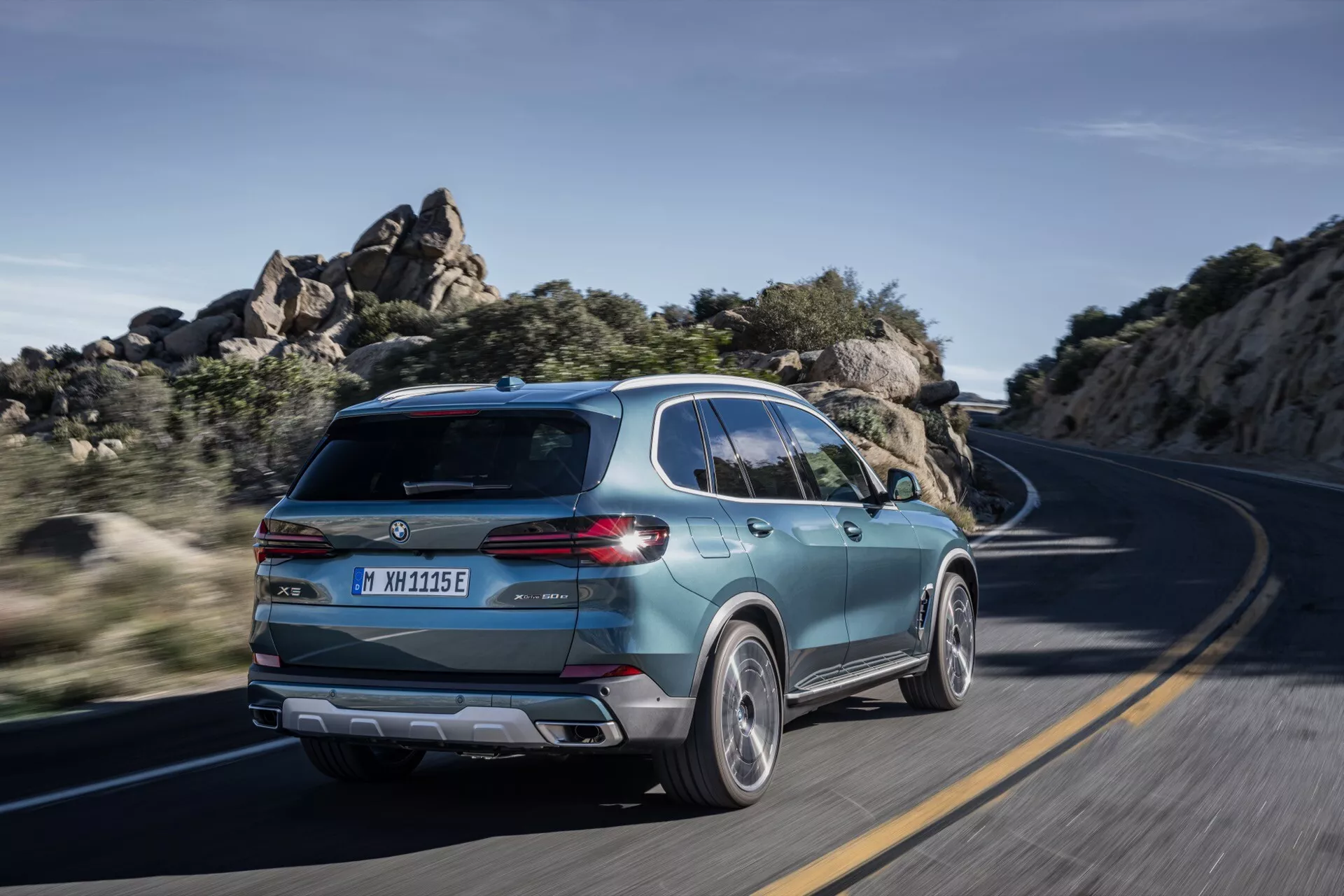 BMW X5 2023 (facelift G05-F95) v pohybe, zachytené z zadného trojštvrťového pohľadu zdôrazňujúceho športový dizajn SUV, moderný zadný kryt s elegantnými zadnými svetlami, dvojfarebné zliatinové kolesá a dôkazy o elektrifikácii na kľukatej ceste za jasného denného svetla