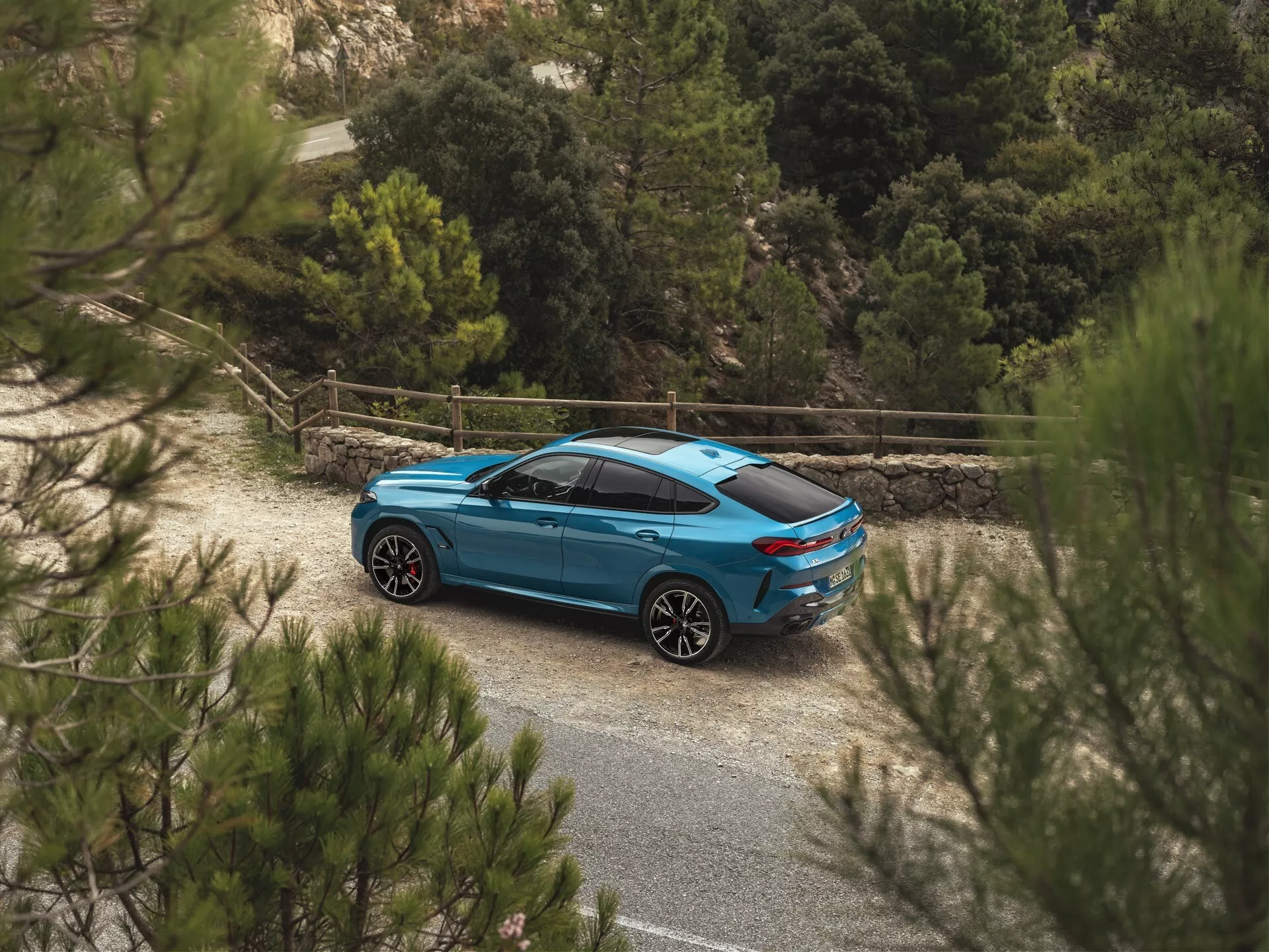 BMW X6 2023 (G06-F96-Facelift) v modrej farbe, sfotografované z vysokého pravého zadného trojštvrťového uhla zdôrazňujúce jej bočný a zadný profil, aerodynamický dizajn, vlastné kolesá a integrované technologické vlastnosti uprostred vidieckeho prostredia s prírodnou vegetáciou
