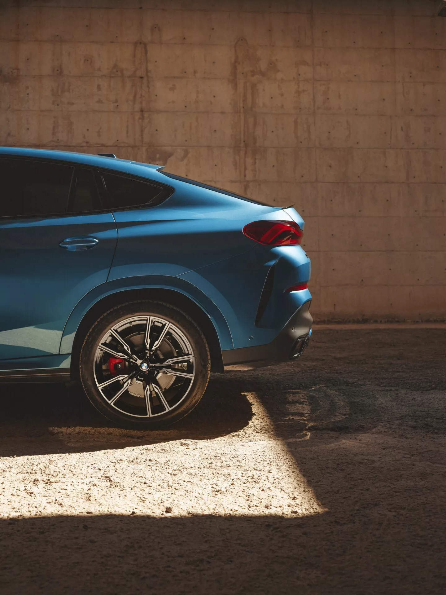 Modré BMW X6 2023 (G06-F96-Facelift) z čiastočného bočného a zadného uhla zdôrazňujúce jeho zadnú pravú štvrtinu s detailným zameraním na viacprvkové zliatinové koleso, červenú výkonnostnú brzdovú strmene a ostrý, uhlový dizajn zadného svetla, zachytené v polotienistom osvetlení vytvárajúce elegantný dynamický kontrast proti betónovému pozadiu.