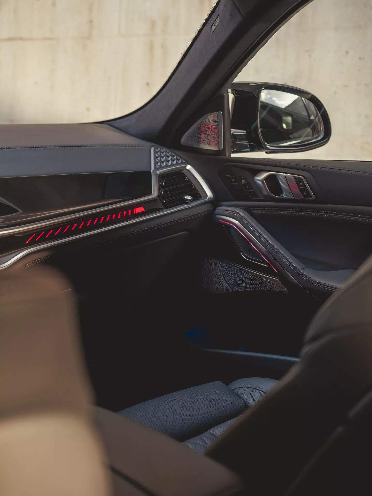Pohľad do interiéru BMW X6 2023 (G06-F96 Facelift) so zameraním na detaily dverí na strane spolujazdca, s ergonomickým dizajnom panela s červeným akcentovým osvetlením, prvotriednymi materiálovými povrchmi a ovládacími prvkami pre elektricky ovládané okná a zrkadlá, doplnené o mriežku reproduktora vysokej vernosti zvukového systému a bezpečnostne orientovaný dizajn s viditeľným bezpečnostným pásom, ilustrujúce technológie pohodlia, pohodlnosti, zvuku, osvetlenia a bezpečnosti.