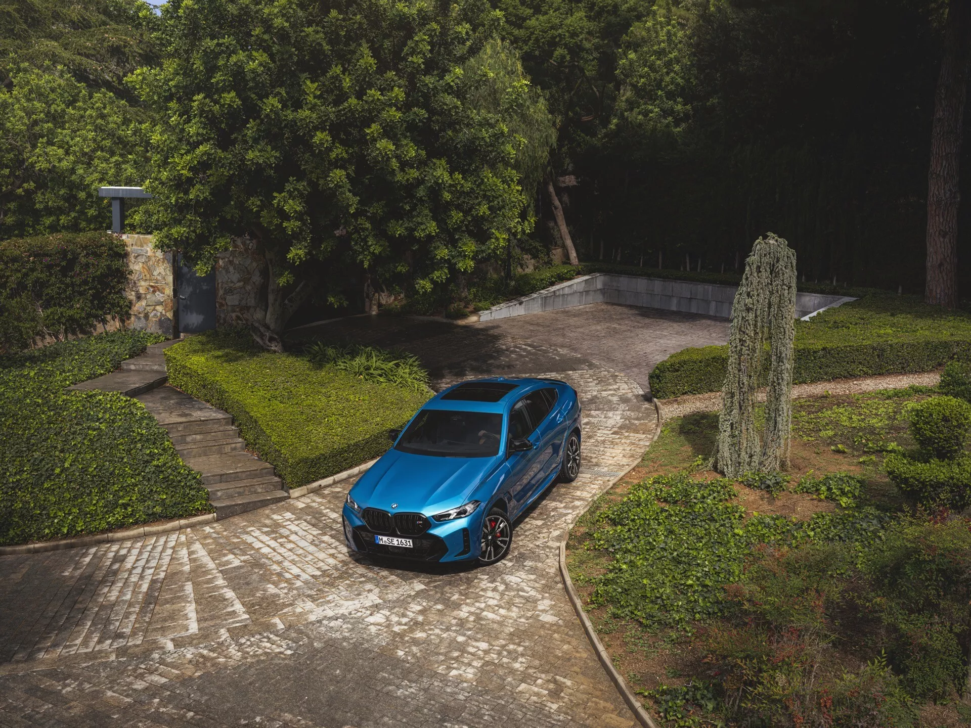 BMW X6 2023 (G06-F96-facelift) zaparkované vonku, ukazuje svoj elegantný metalický modrý finish a moderný dizajn, vrátane veľkých zliatinových kolies a charakteristickej sklonenej strechy medzi prostredím kamenitých chodníkov a bujnej zelene pod mäkkým slnečným svetlom
