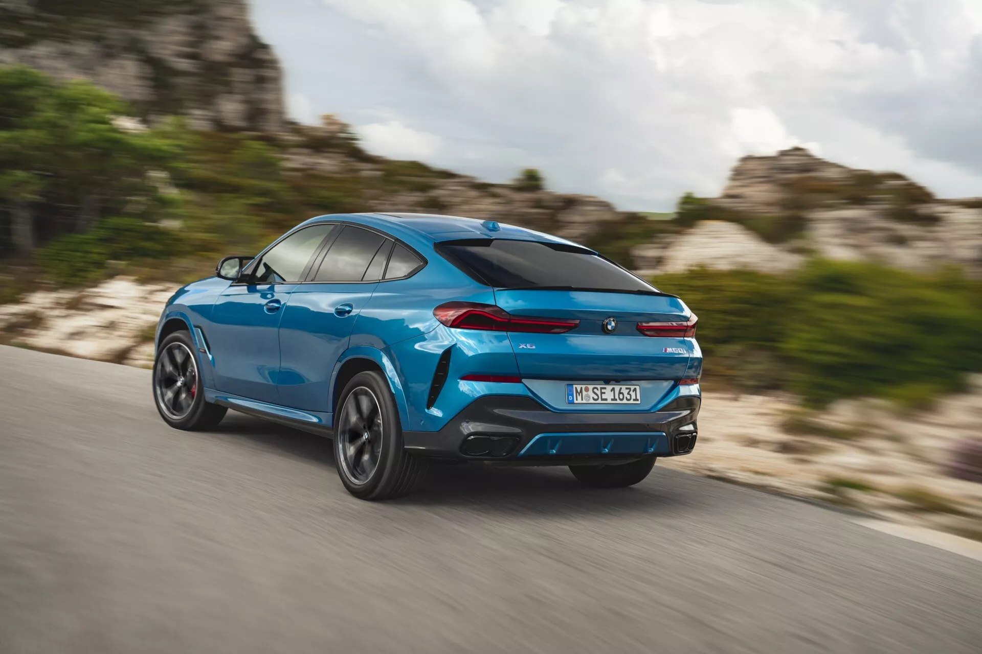 BMW X6 2023 (G06-F96-Facelift) vo výraznom modrom odtieni, zachytené v dynamickom zobrazení zo zadného trojštvrtečného uhla ukazujúci bočný profil, otáčajúce sa kolesá a elegantný dizajn SUV typu kupé s výkonnostnými prvkami ako sú dvojité výfuky a zadný spojler pod efektom rozmazania pri pohybe na diaľnici.