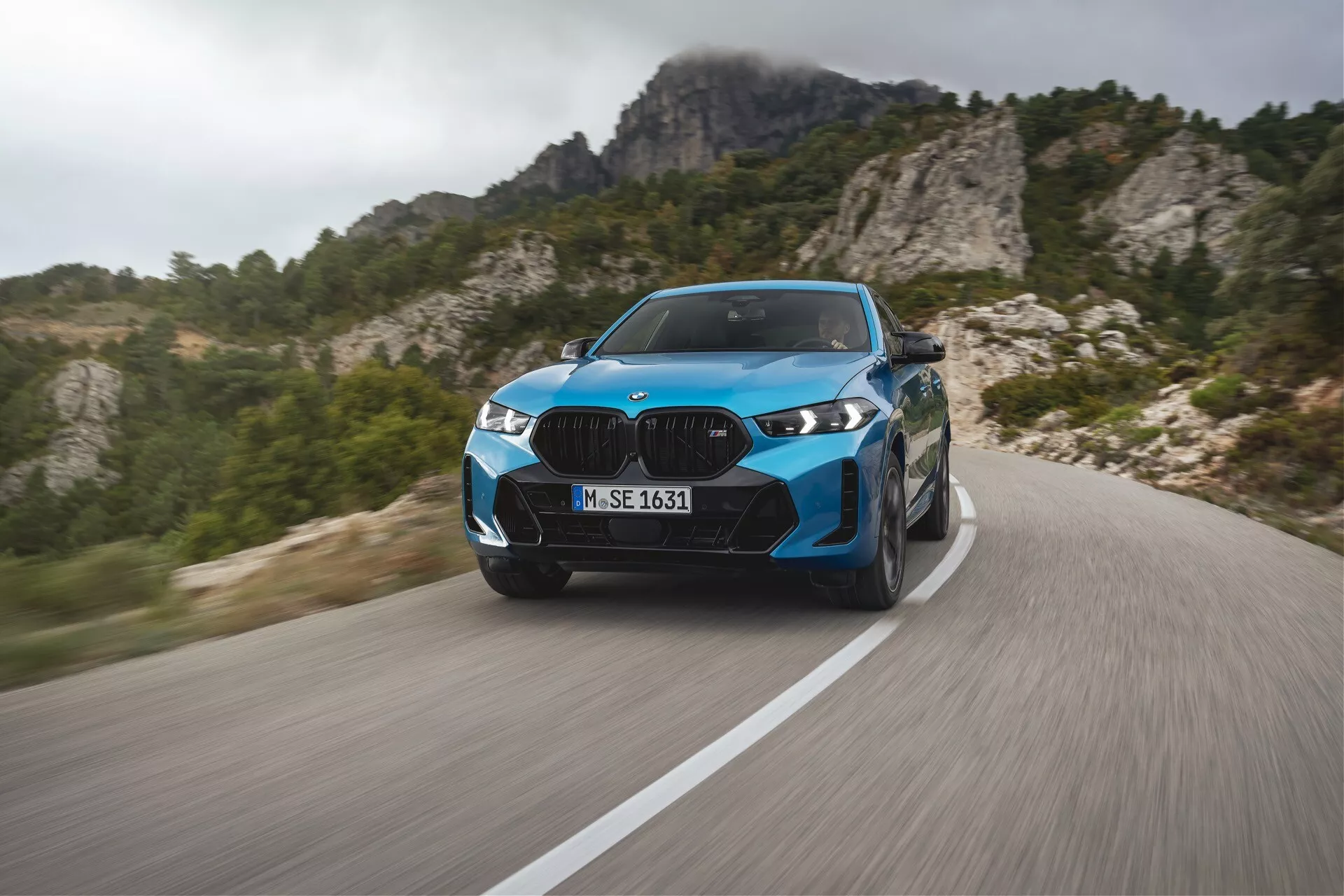 BMW X6 2023 (G06-F96-facelift) vo výraznej modrej farbe, predvádzajúce moderné luxusné výkony na kľukatej horskej ceste s dynamickým rozmazaním pohybu, pokročilými LED svetlometmi, štíhlymi dizajnovými prvkami a športovými kolesami na pozadí drsnej horskej krajiny.
