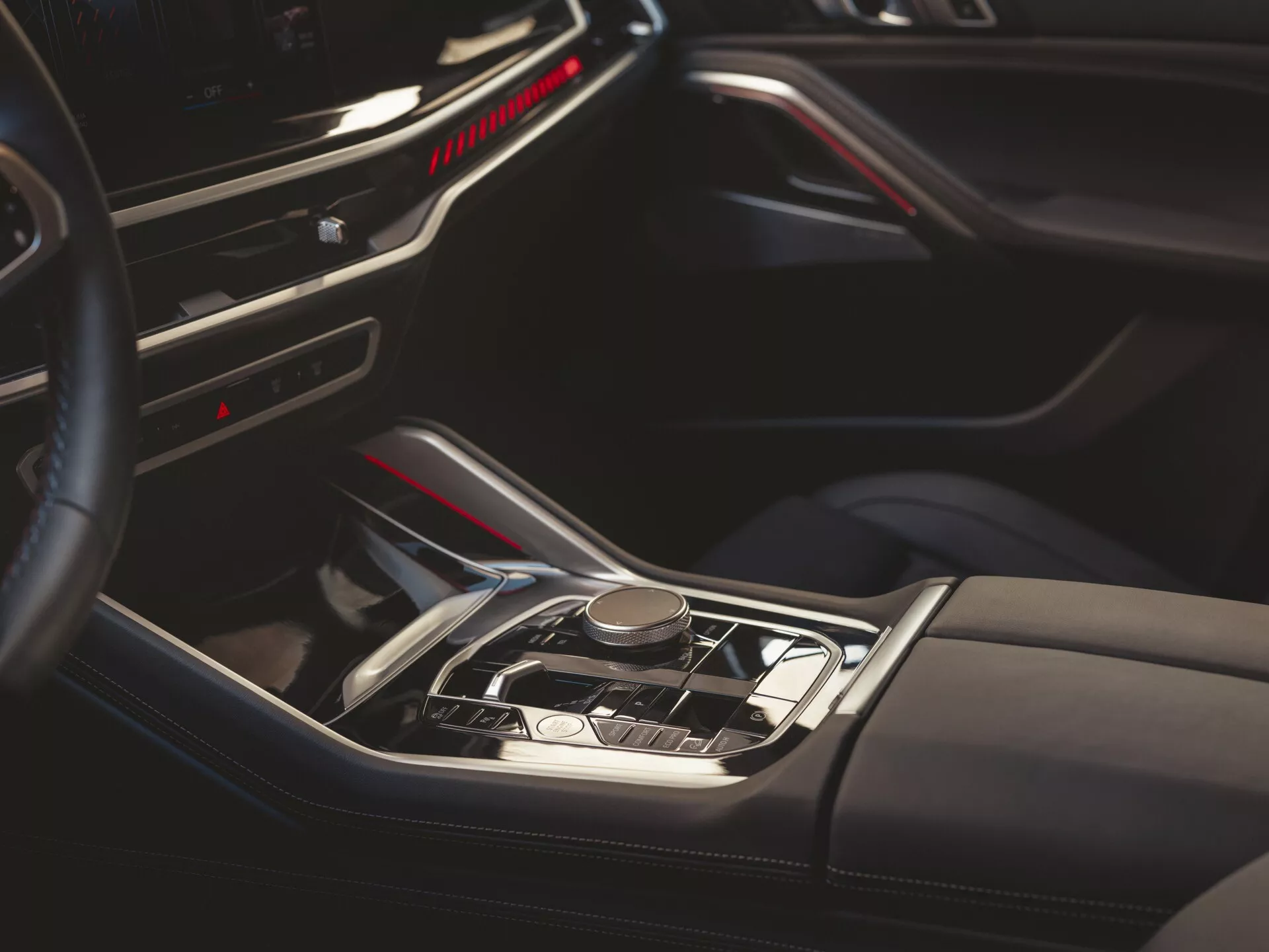 Detailný pohľad na interiér BMW X6 2023 (G06-F96-Facelift) predstavujúci automatickú radicu páku, luxusnú stredovú konzolu s imitáciou dreva, kovovými akcentmi, červeným ambientným osvetlením, koženými detailmi a čiastočným výhľadom na prístrojovú dosku a dvere vodiča