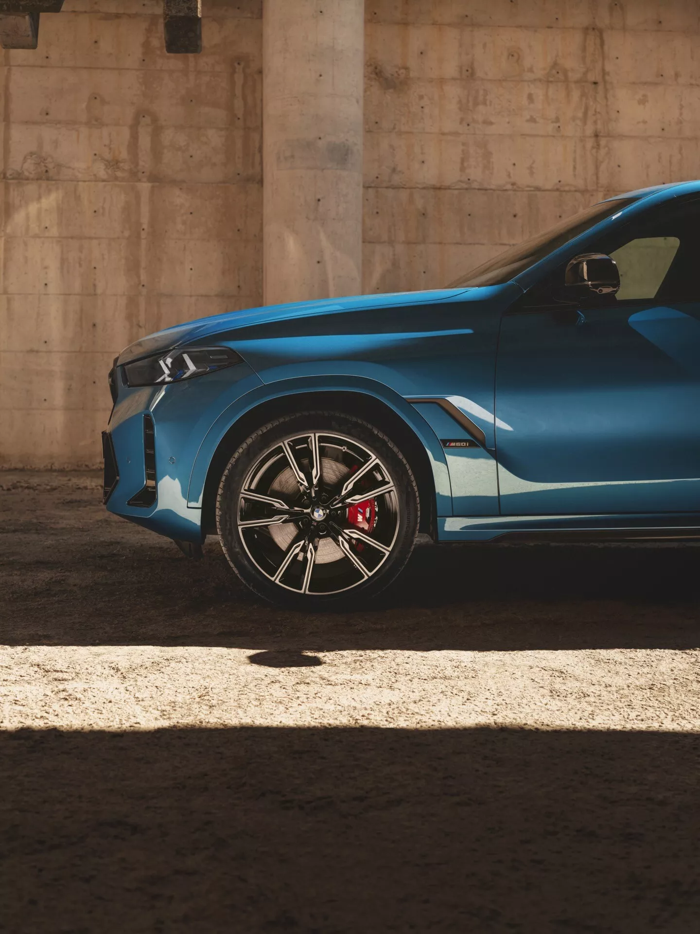 Čiastočný pohľad na pravú prednú štvrtinu BMW X6 2023 (G06-F96-Facelift) ukazuje jeho moderný agresívny dizajn zdôraznený ostrou charakterovou líniou, športovým zliatinovým kolesom s červeným vysokovýkonným brzdovým strmcom a nízkoprofilovou pneumatikou, pod dramatickým slnečným svetlom vytvárajúcim vysoký kontrast tieňov a zvýraznení.