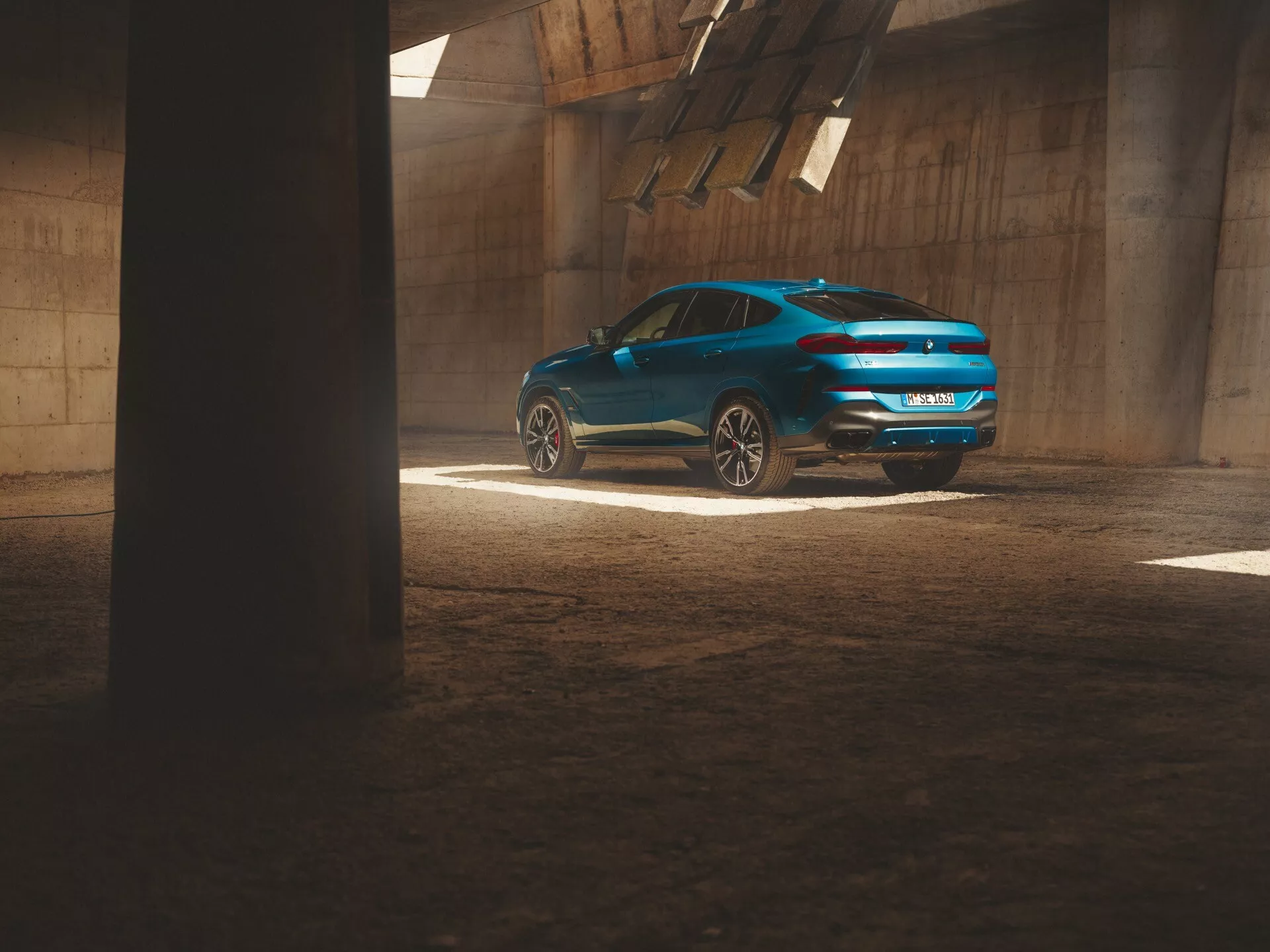BMW X6 2023 (G06-F96 Facelift) v metalickom modrej, ukazuje z trojštvrtečný pohľad zozadu v priemyselnom prostredí, zdôrazňuje športový dizajn, LED zadné svetlá a dvojitý výfukový systém pod dynamickým osvetlením