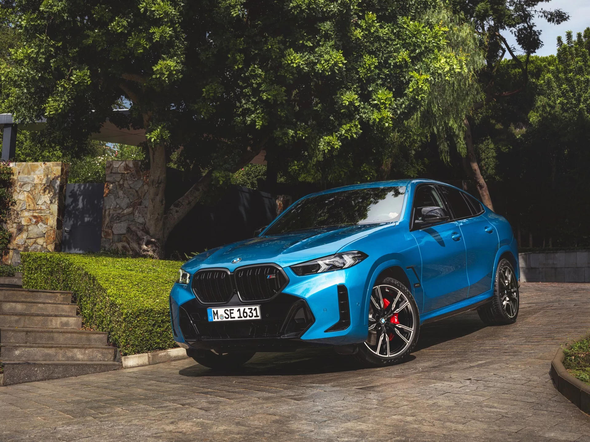 BMW X6 2023 (G06-F96-Facelift) v modrej farbe, zaparkované pod uhlom ukazujúce svoju prednú časť, obličkovitý dizajn mriežky, LED alebo laserové predné svetlá, športové zliatinové kolesá so červenými brzdovými strmienmi, kontúrované línie kapoty a aerodynamické bočné zrkadlá s integrovanými smerovkami, zachytené v prírodnom dennom svetle so zeleňou a architektonickými schodmi na pozadí