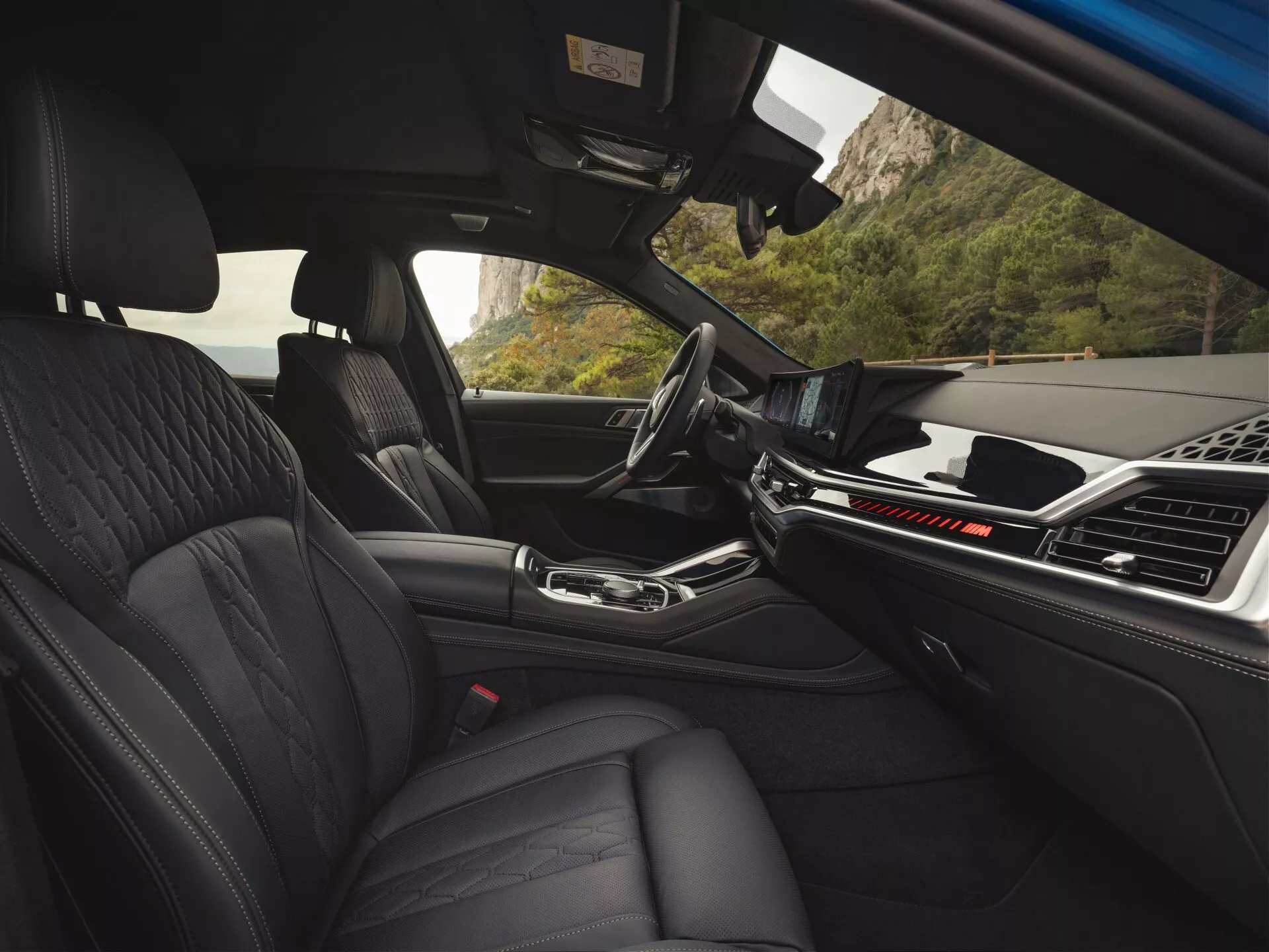 Interiér BMW X6 2023 (G06-F96 Facelift) pohľad zo zadného sedadla ukazujúci prednú palubnú dosku, predné sedadlá z prémiovej kože s prešívaným dizajnom, centrálnu lakťovú opierku a pokročilý dotykový informačný systém, obalený v modernom, minimalistickom estetickom vzhľade naznačujúcom luxus a špičkovú automobilovú technológiu