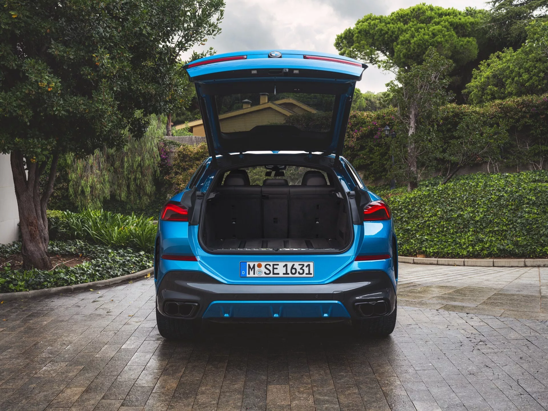 Zadný pohľad na BMW X6 2023 (G06-F96-facelift) na drevenej terase s otvoreným kufrom bez nákladu, ukazujúci kapacitu batožinového priestoru a ľahký prístup, na pozadí zelenej vegetácie v rezidenčnom alebo parkovom prostredí, vrátane funkcií ako je elektrické otváranie kufra, možná asistencia pri parkovaní alebo systém kamery dozadu, s mäkkým, rozptýleným osvetlením zdôrazňujúcim detaily dizajnu vozidla