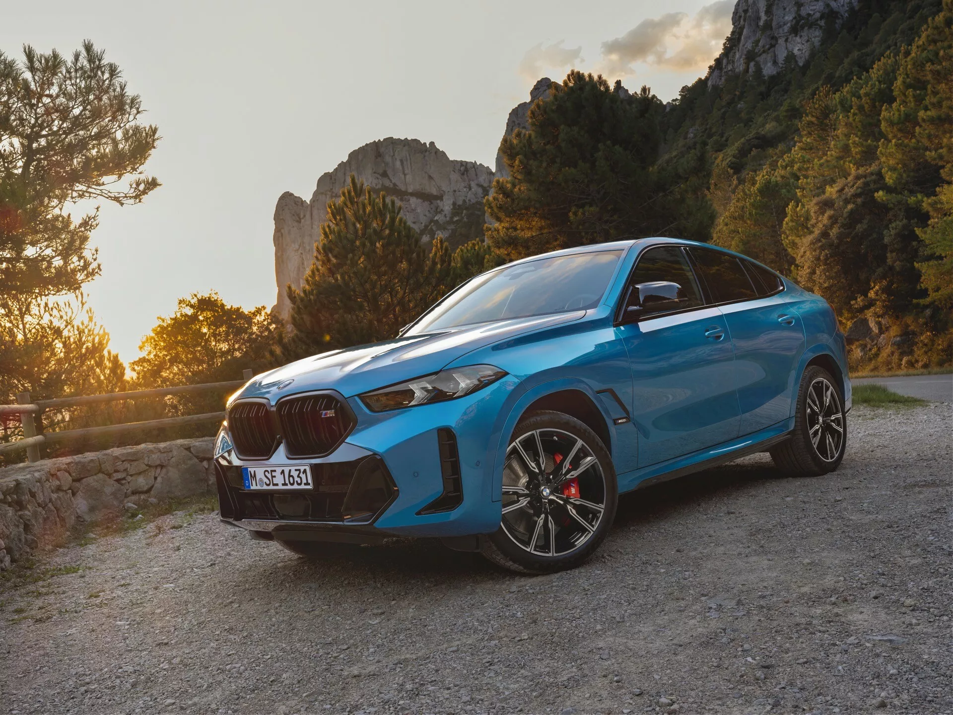 BMW X6 2023 (G06-F96-Facelift) v modrej farbe, predstavujúce športový SUV dizajn zachytený počas zlatého hodiny s pohľadom z troch štvrtín z predu, ukazujúci charakteristickú veľkú obličkovú mriežku, štíhle LED svetlomety a predný nárazník orientovaný na výkon s veľkými vzduchovými vstupmi, na pozadí pokojnej horskej cesty.