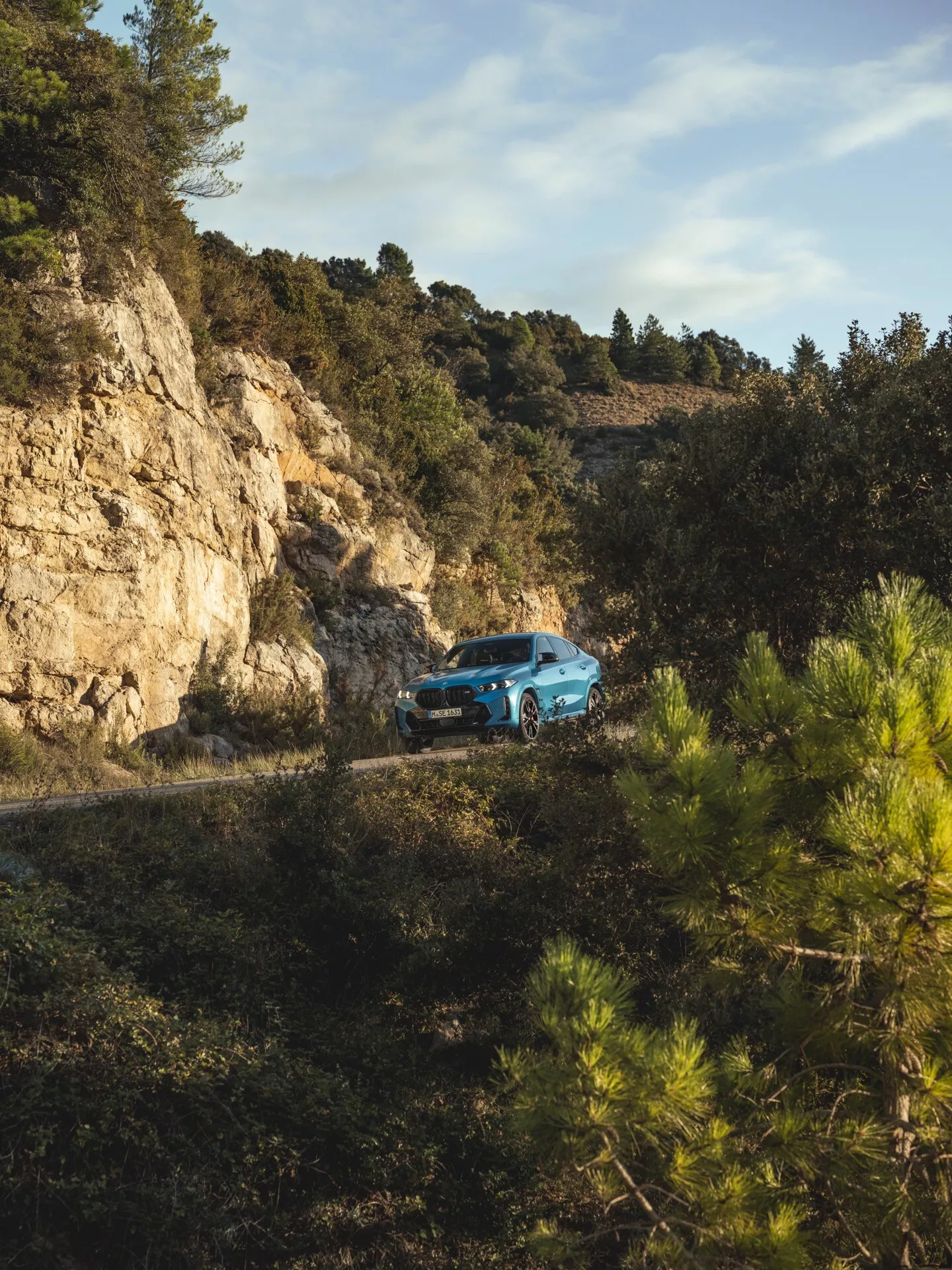 BMW X6 2023 (facelift G06-F96) vo výhľade z troch štvrtín vpredu na zákrute horskej cesty obklopenej lesom, demonštruje schopnosť off-road jazdy a moderný dizajn