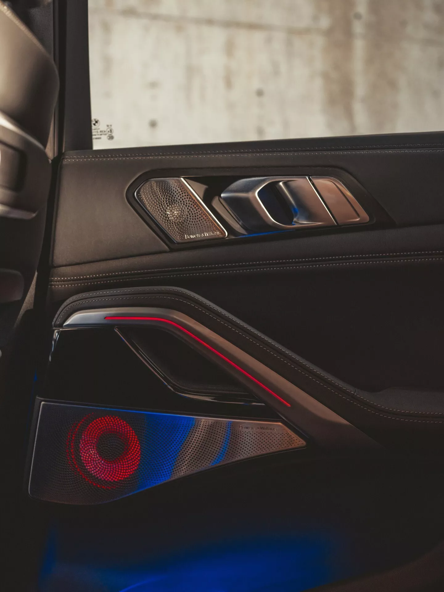 Detail interiéru BMW X6 2023 (G06-F96-Facelift) so zameraním na dvere vodiča s chrómovou kľučkou, dotykovými ovládačmi okien, mriežkou prémiového reproduktora, vreckom na dverách s červeným ambientným osvetlením a výrazným náladovým osvetlením