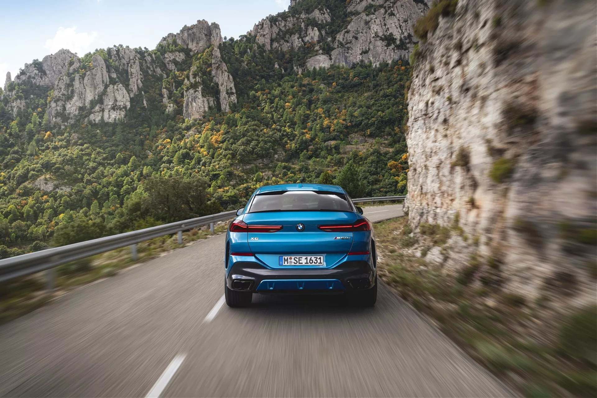 BMW X6 2023 (G06-F96-Facelift) vo vibrantom modrom so zapálenými LED zadnými svetlami, športovým aerodynamickým profilom so spoilerom na streche, dvojitým výfukovým systémom, na horskej ceste ukazujúcom rozostrenie pohybu na zdôraznenie výkonu a pohybu