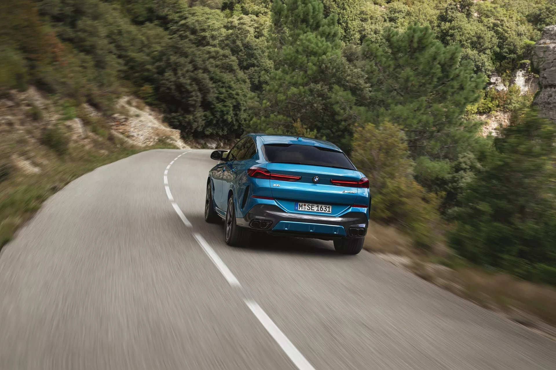 Dynamický zadný pohľad na modré BMW X6 2023 (G06-F96-Facelift) so športovým dizajnom a modernými zadnými svetlami na kľukatej ceste uprostred prírodnej krajiny