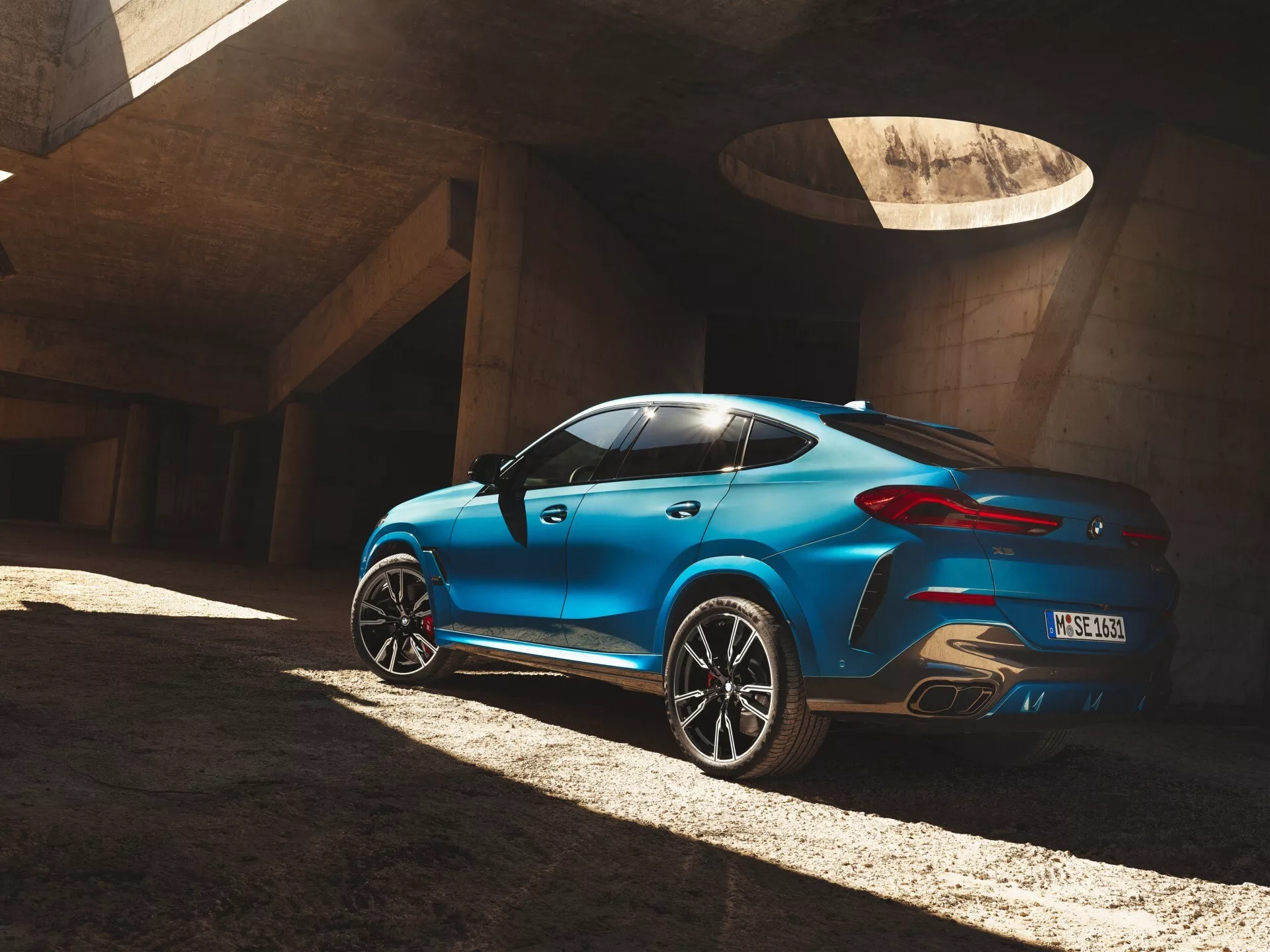 BMW X6 2023 (G06 F96 facelift) v modrej farbe, zachytené z uhla tri štvrtiny zozadu, zvýrazňujúce dizajnové línie vozidla, siluetu, tvar zadného okna, oblúky zadných kolies a zadné svetlá, zaparkované na betóne pod kontrastným slnečným svetlom.
