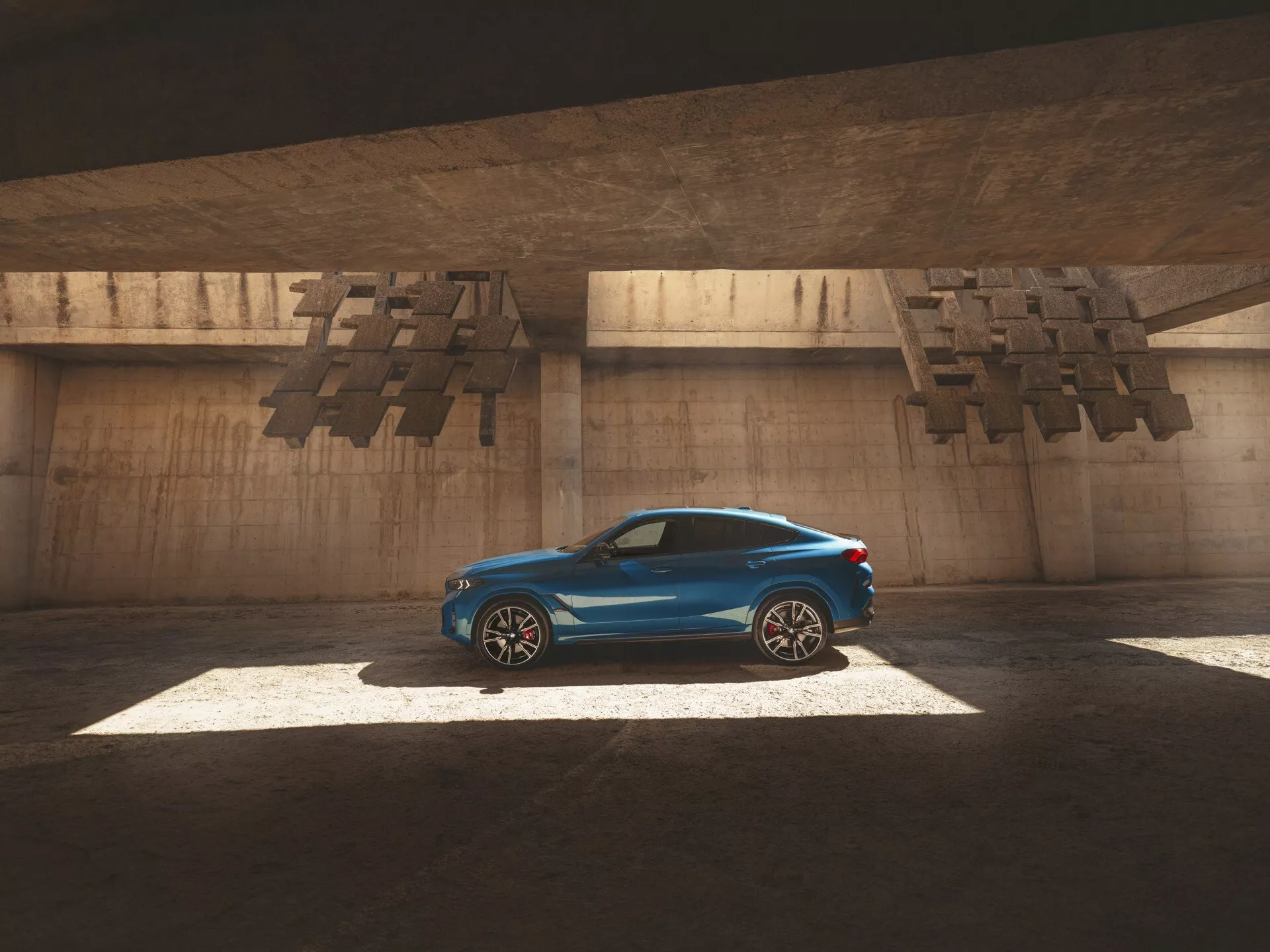 Kupé štýlu auto BMW X6 2023 (G06-F96 Facelift) v lesklom modrom odtieni, osvetlené slnkom cez otvor nad autou, zachytávajúce jeho štíhly bočný profil, aerodynamické línie a luxusné prvky ako sú zliatinové kolesá a strešné okno