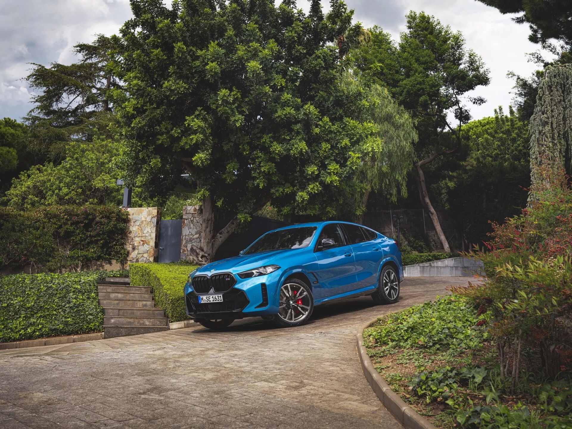 BMW X6 2023 (G06-F96-Facelift) v modrej farbe, trojštvrťový pohľad spredu, zaparkované na príjazdovej ceste s okolitou zeleňou, zobrazujúce predný nárazník, mriežku, kapotu, čelné sklo, profil vodiča, LED svetlomety, športové kolesá s červenými brzdovými strmienkami a aerodynamický dizajn