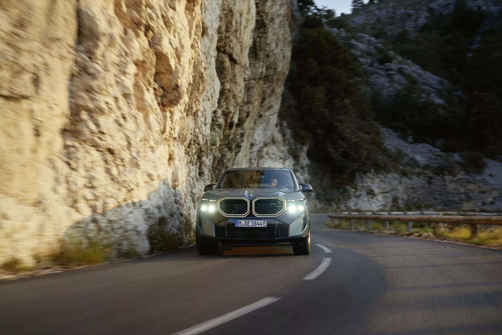BMW XM (G09) z roku 2023 jazdiace po zákrutej horskej ceste v súmraku, s LED svetlometmi, osvetleným palubným doskou a moderným modro-šedým lakom