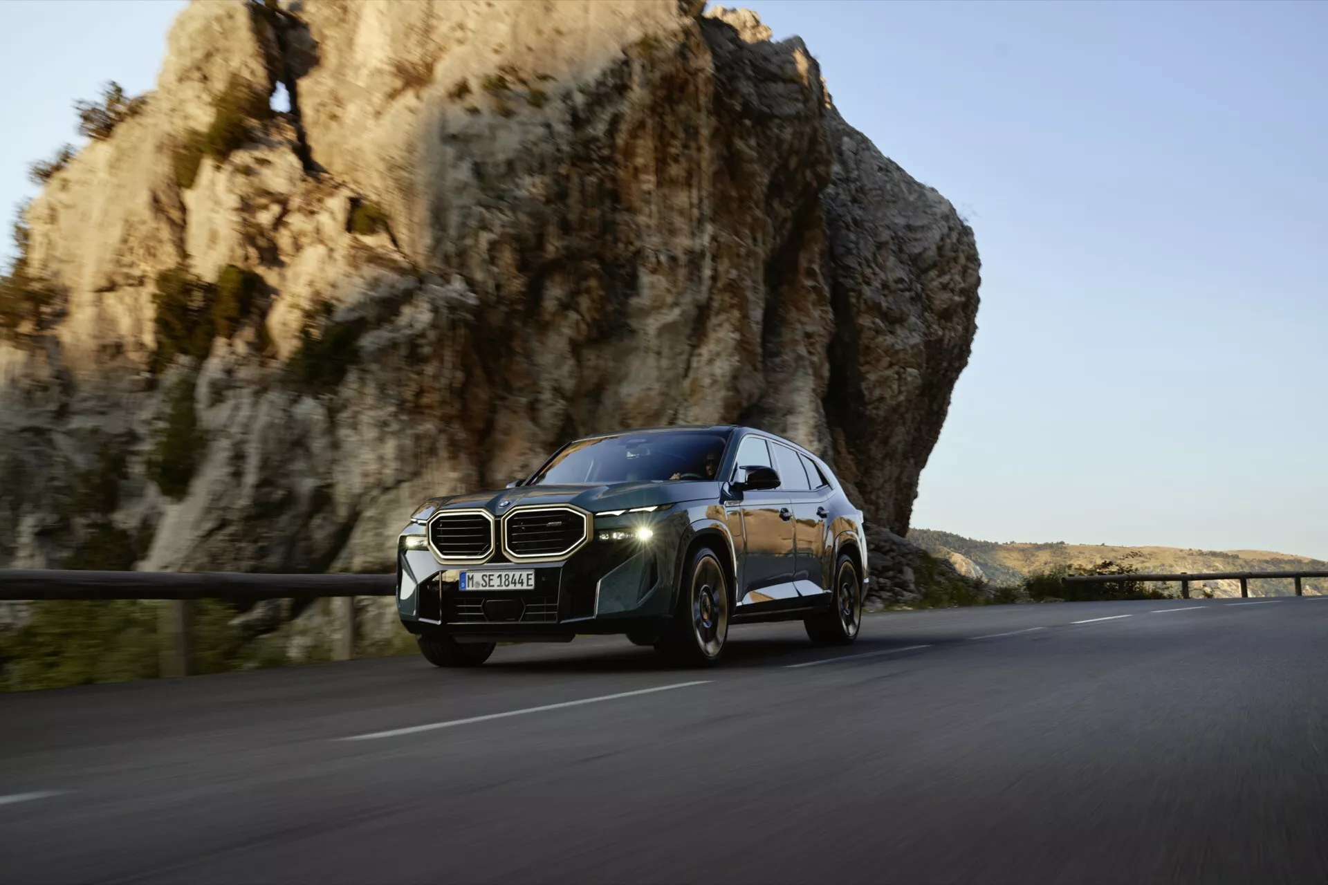 Luxusné SUV BMW XM (G09) z roku 2023 v pohybe na malebnej trase s charakteristickou obličkovou mriežkou, modernými LED svetlometmi a aerodynamickým dizajnom na pozadí skalného útesu a teplého slnečného svetla.