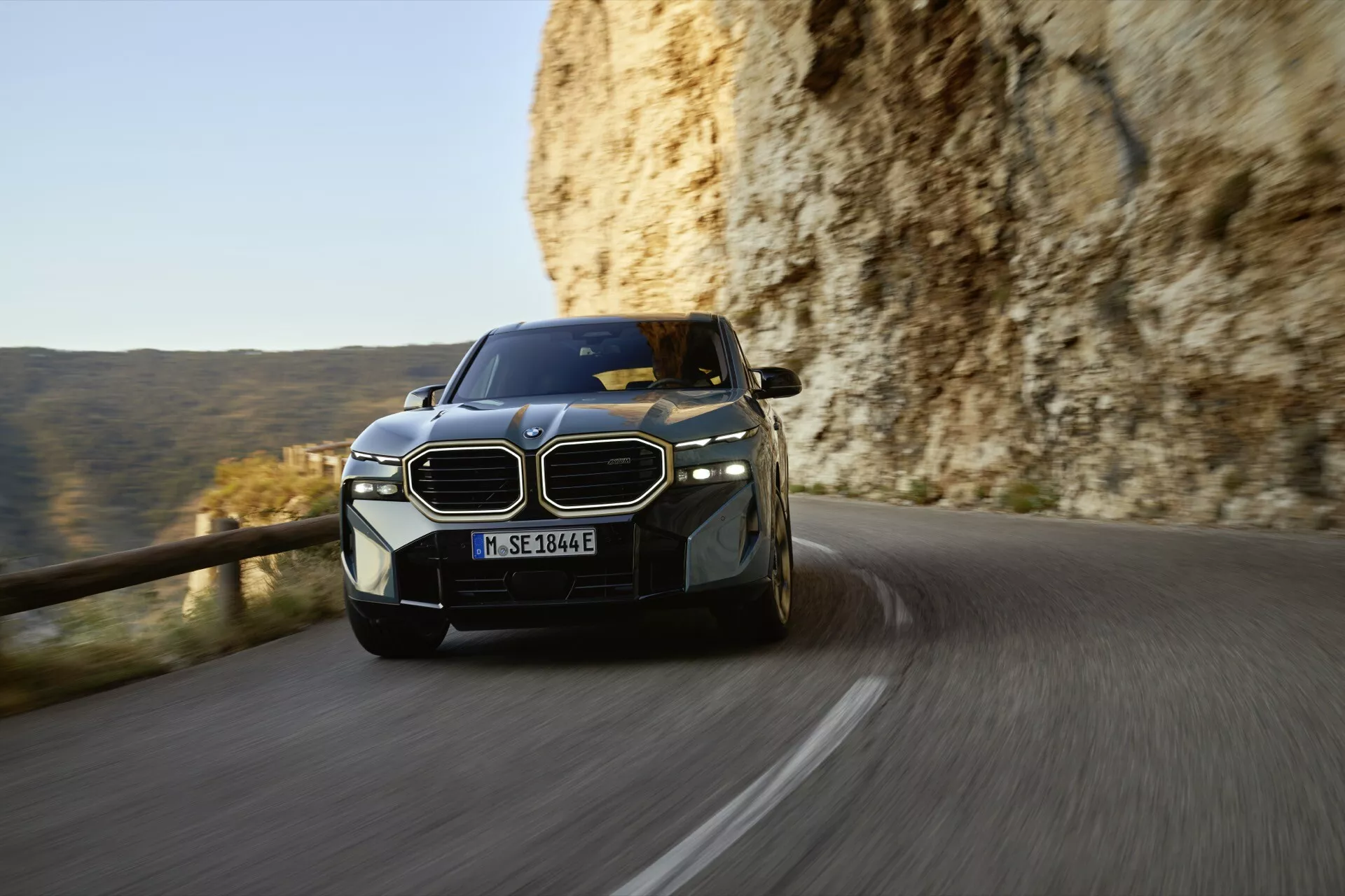 BMW XM (G09) z roku 2023 v pohybe na horskej ceste, ukazuje svoj dynamický postoj, LED svetlomety a charakteristickú obličkovú mriežku z nízkeho uhla pohľadu spredu v neskoré popoludnie