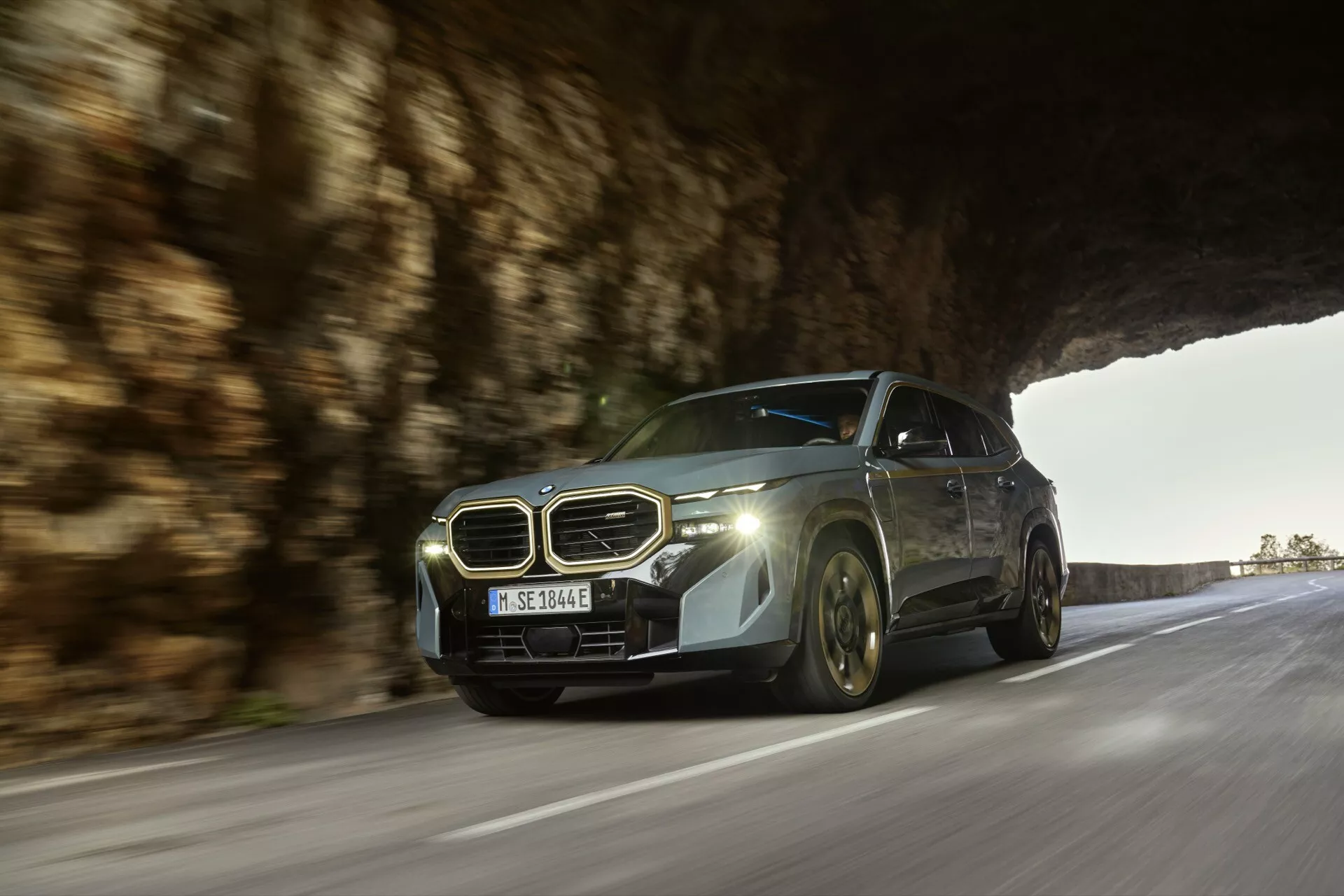 Dynamický záber z nízkeho uhla BMW XM 2023 (G09) v pohybe na ceste, zdôrazňujúci jeho moderný luxusný dizajn, charakteristickú obličkovitú mriežku, emblém, pokročilé LED svetlomety a elegantnú aerodynamickú karosériu s kovovým povrchom pri prirodzenom osvetlení