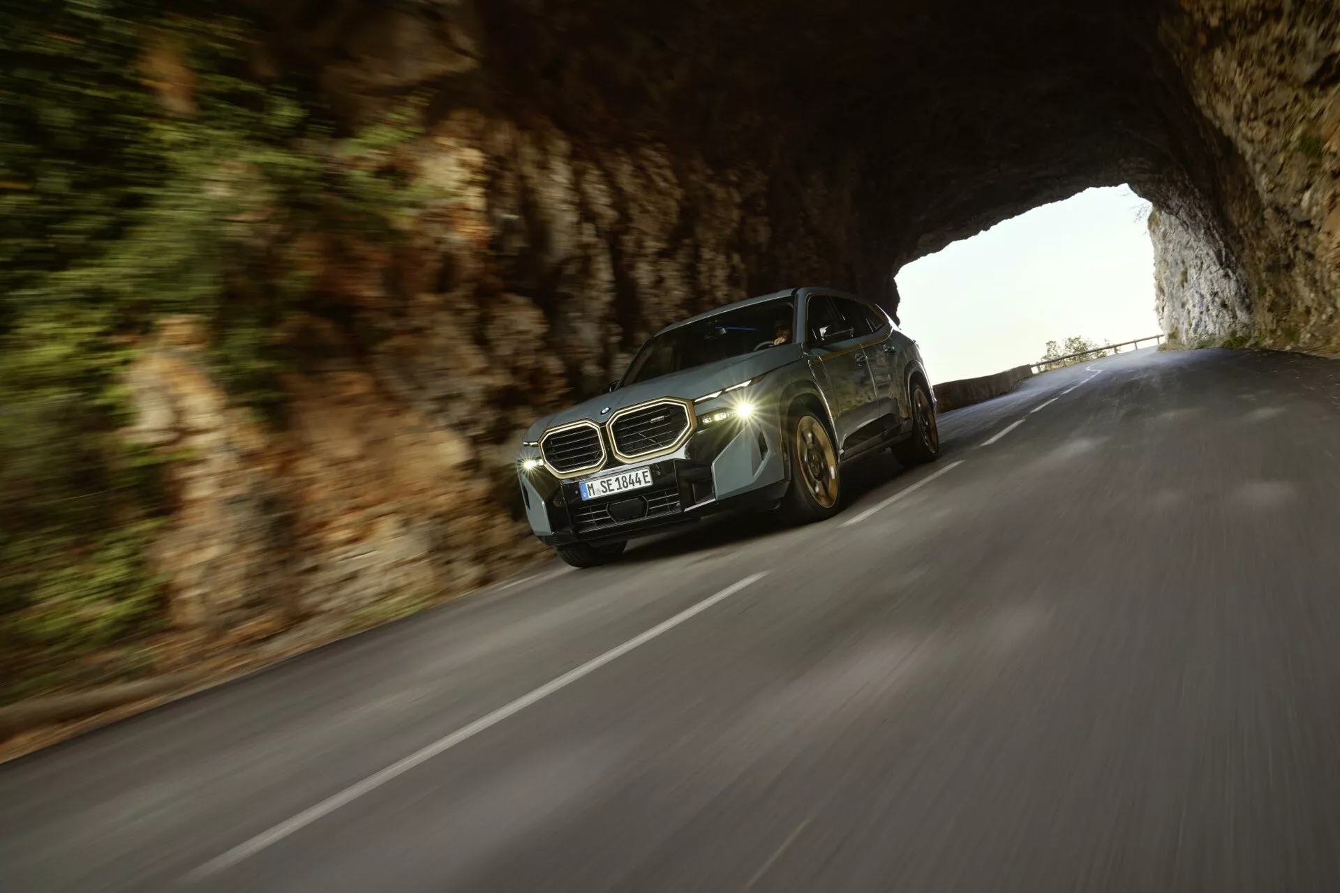 Dynamický pohyb BMW XM (G09) z roku 2023, predvádzajúci osvetlené LED svetlomety a modernú mriežku chladiča, pri výjazde z kamenného tunela na spevnenú cestu