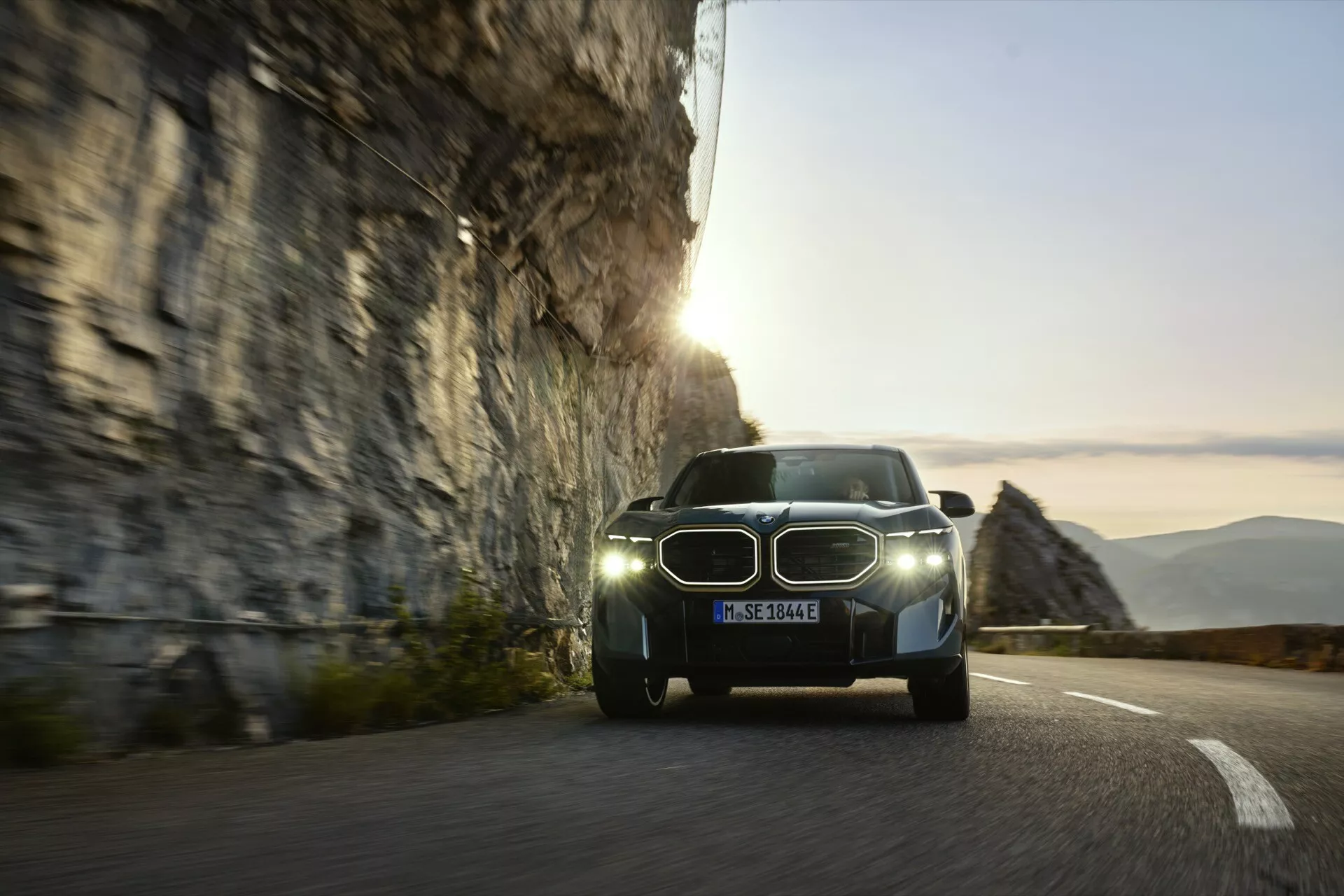 BMW XM (G09) roka 2023 zobrazené na zákrutej horskej ceste pri východe alebo západe slnka, s charakteristickými LED svetlometmi, ikonickou obličkovou mriežkou, potenciálnym elektrickým pohonom so senzormi pre asistenciu vodiča a dynamickým rozmazaním naznačujúcim pohyb.