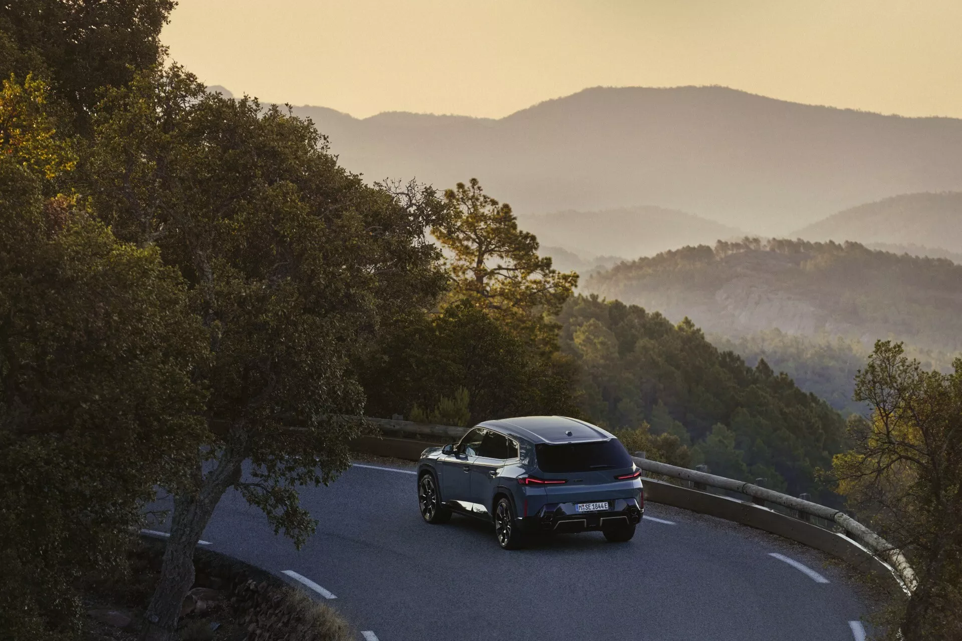 BMW XM (G09) z roku 2023 zobrazené z trojštvrtečného zadného uhla na horskej ceste počas východu alebo západu slnka, predvádzajúce jeho elegantný dizajn, LED zadné svetlá a zliatinové kolesá