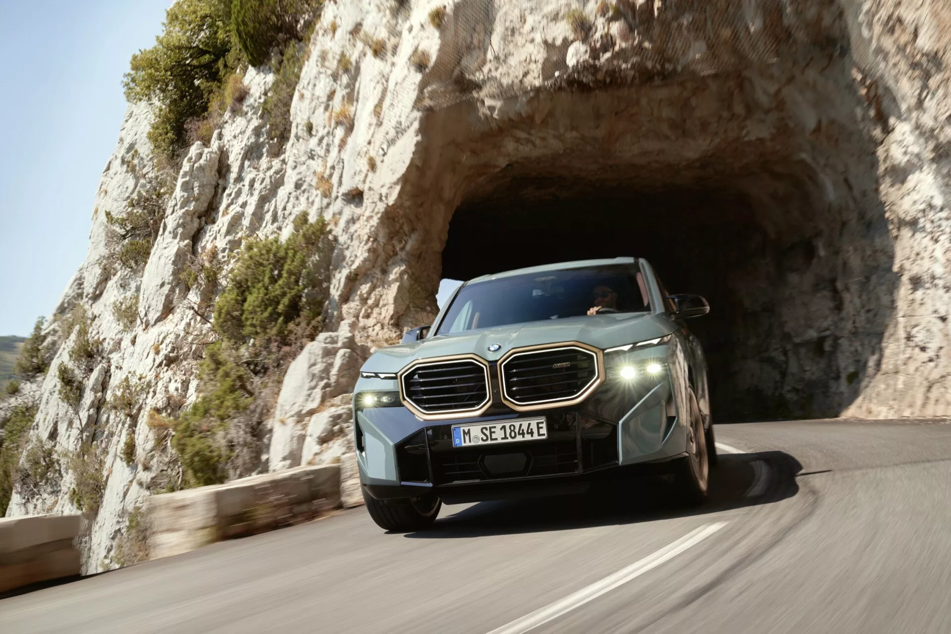 Luxusné SUV 2023 zachytené v pohybe, ako vychádza z prírodného skalného oblúka alebo tunela, predvádzajúce svoj aerodynamický dizajn a pokročilé technologické funkcie ako LED svetlomety a senzory asistencie vodiča, s európskou poznávacou značkou, na pozadí scénickej pobrežnej alebo horskej krajiny.