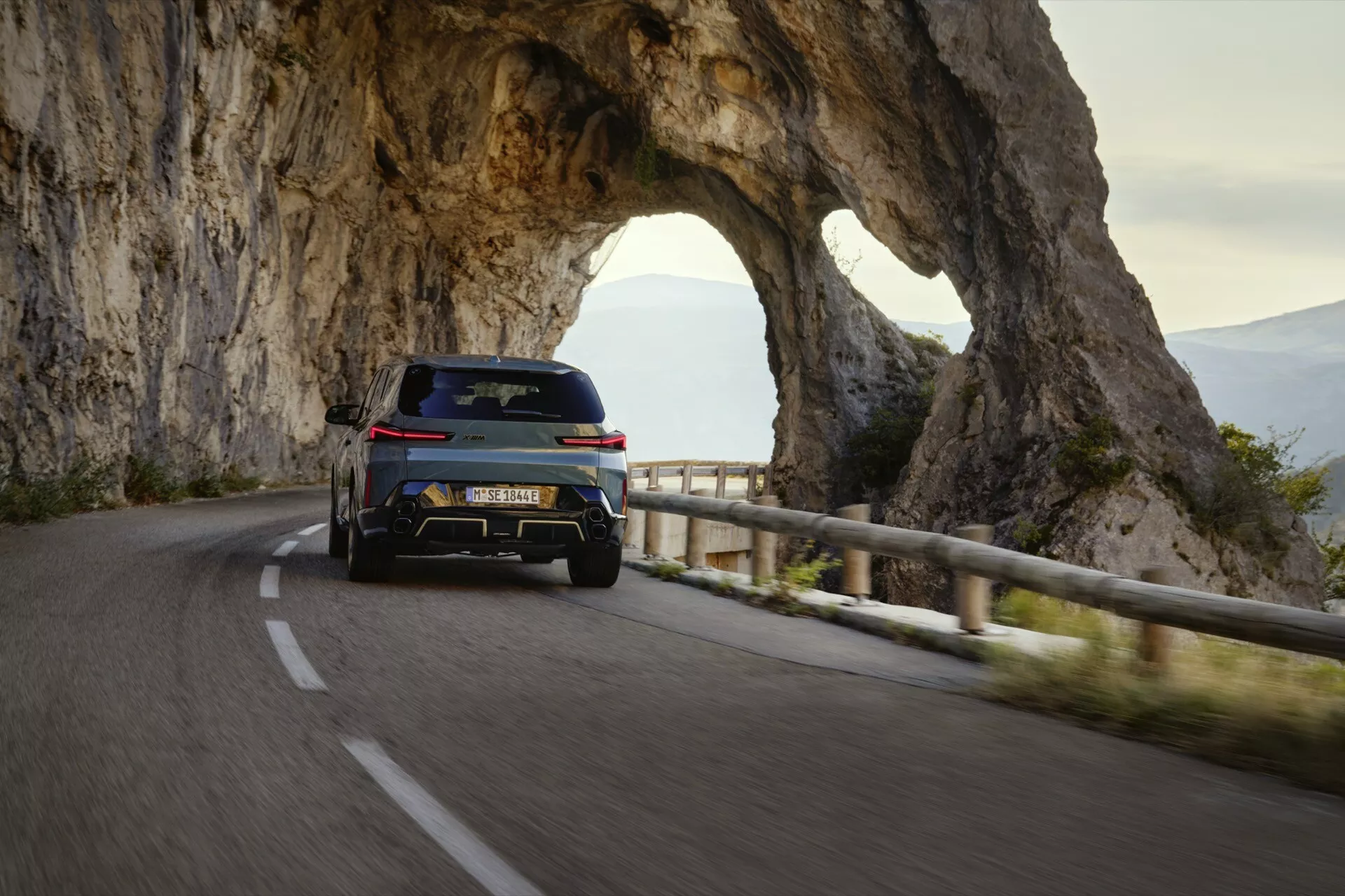 BMW XM (G09) z roku 2023 zachytené z uhla troch štvrtín zozadu na horskej ceste, zdôrazňujúce jeho duálny výfukový systém, štýlové zadné svetlá, zadný difúzor pre aerodynamiku, zdvihnutý dizajn SUV, veľké zliatinové kolesá, zadný spojler, tónované zadné okná a športový bočný profil v večernom osvetlení
