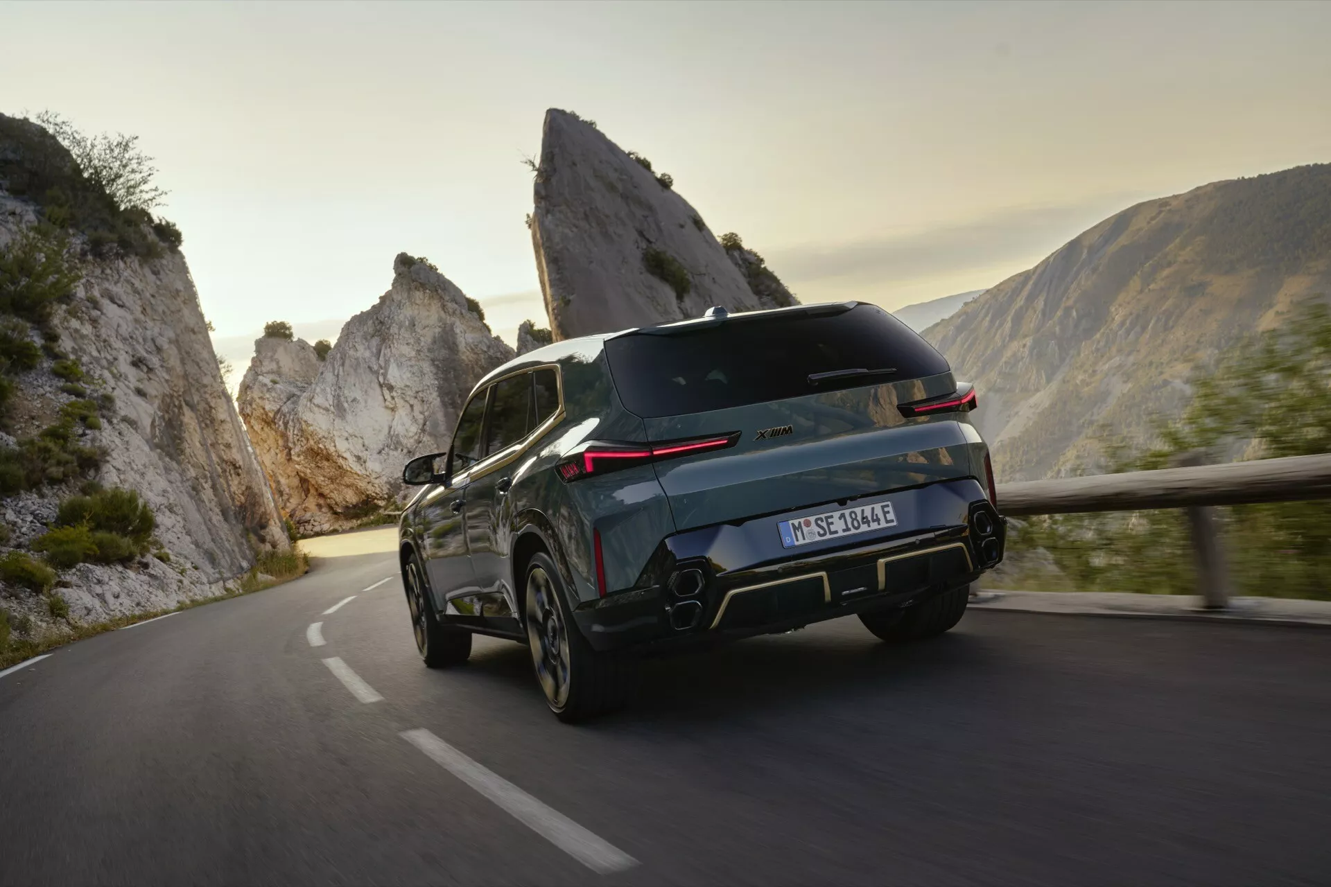 Luxusné SUV BMW XM (G09) z roku 2023 v pohybe na horskej ceste, zachytené z zadného trojštvrtečného uhla zdôrazňujúceho jeho športovú siluetu, elegantné zadné LED svetlá, aerodynamické línie a pokročilé bezpečnostné funkcie proti dramatickej prírodnej krajine.