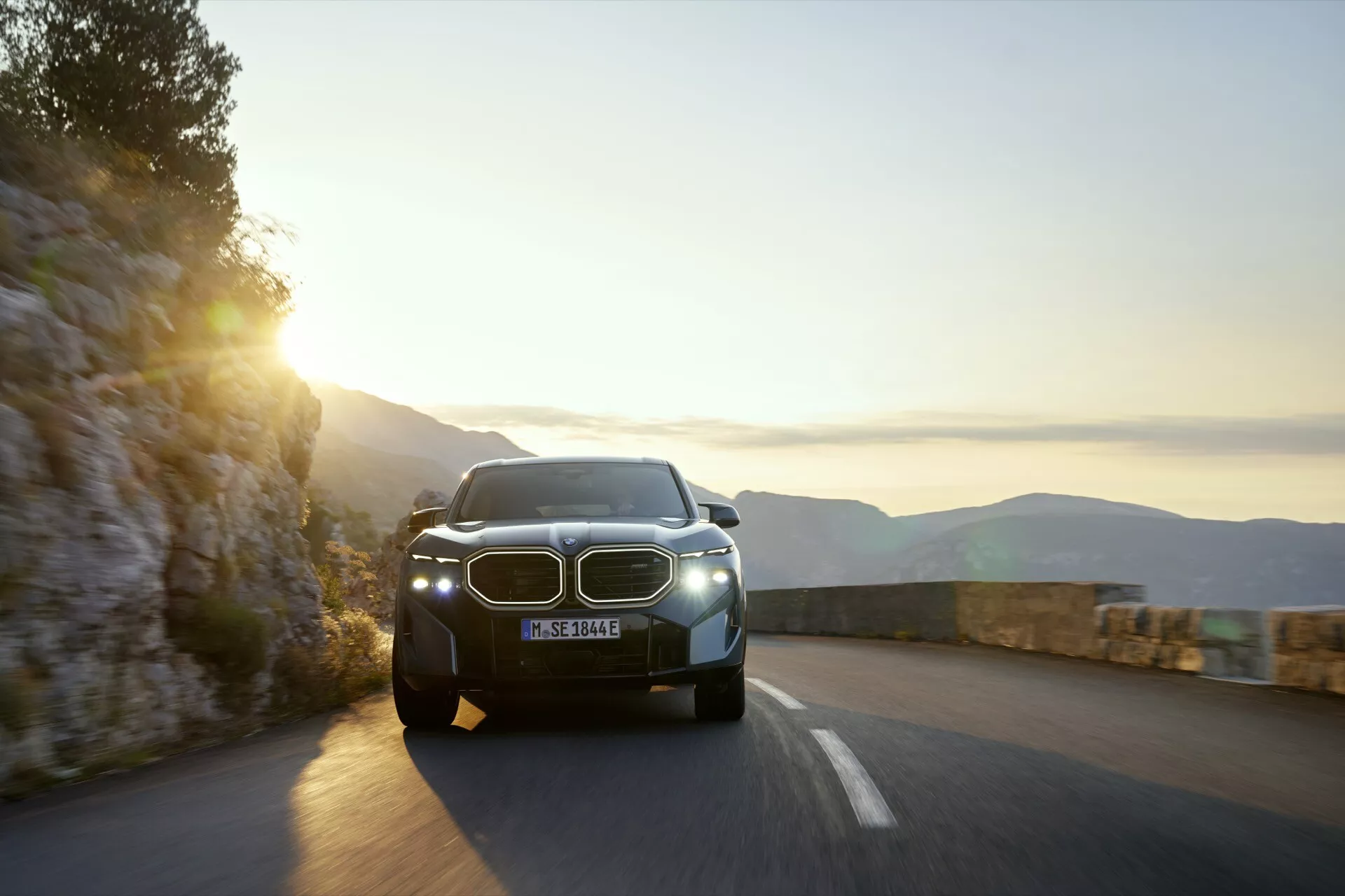 Moderné SUV BMW XM (G09) z roku 2023 idúce po horskej ceste na svitanie alebo súmraku s osvetlenými LED svetlometmi a viditeľnou charakteristickou obličkovitou mriežkou