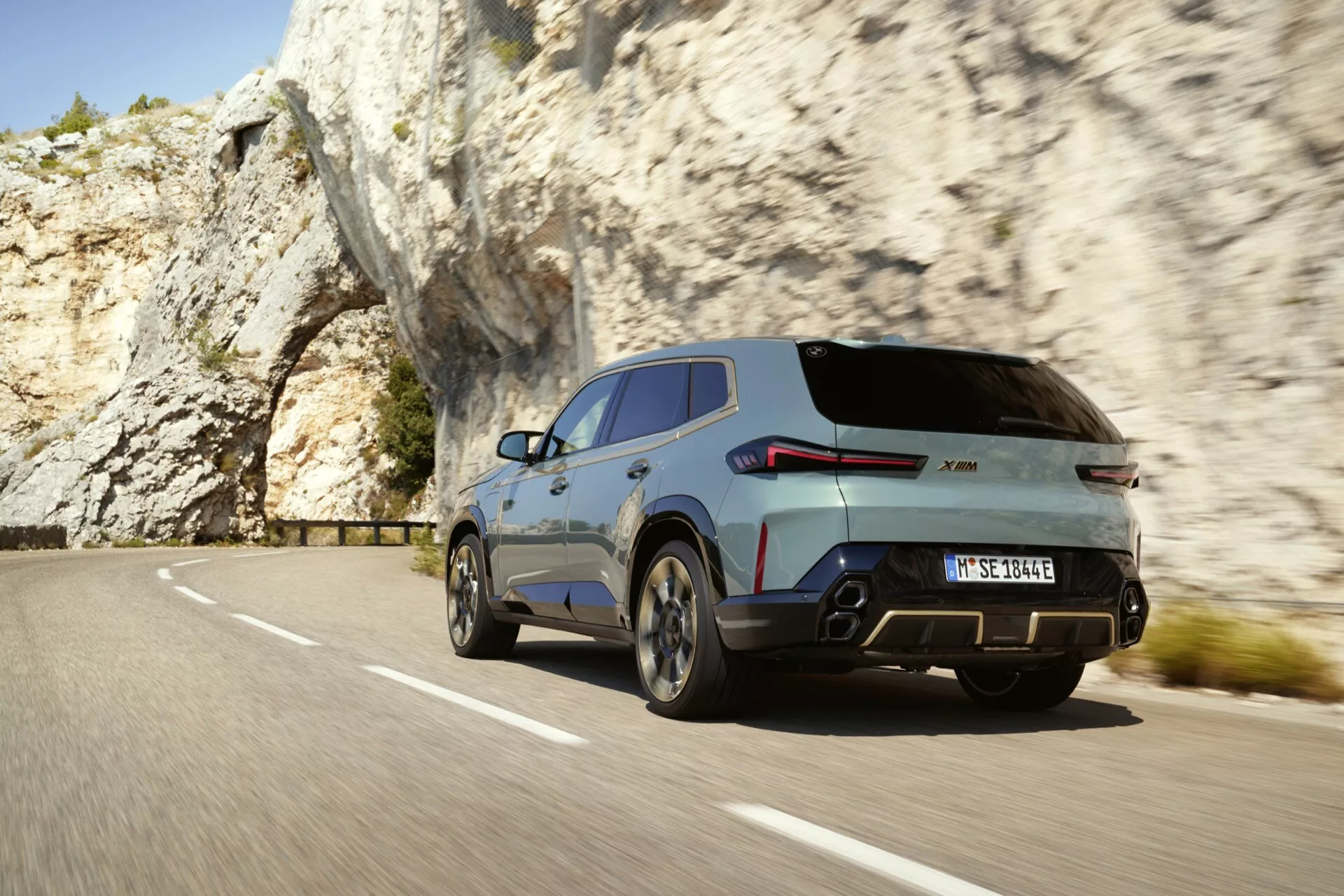Dynamický zadný trojštvrťový pohľad na moderné SUV, pravdepodobne BMW XM (G09) z roku 2023, pohybujúce sa po horskej ceste a zobrazujúce pravé zadné koleso, zadné svetlo, veko batožinového priestoru a časť pravého bočného panela smerom k predným dverám, s viditeľným rozostrením pohybu, dvojitým výfukovým systémom, strešným spojlerom a veľkými zliatinovými kolesami s metalickým povrchom, obklopeným prírodnými skalnými formáciami.