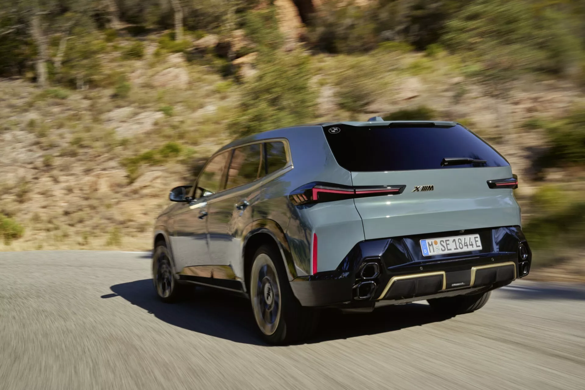 Moderné SUV BMW XM (G09) z roku 2023 v pohybe na spevnenej ceste, s aerodynamickým športovým dizajnom, LED zadnými svetlami, karosériou v dvoch tónoch, odrazovými parkovacími senzormi, polygonálnym duálnym výfukom, zliatinovými kolesami a možným označením elektrického/hybridného vozidla na pozadí vidieckej / horskej krajiny