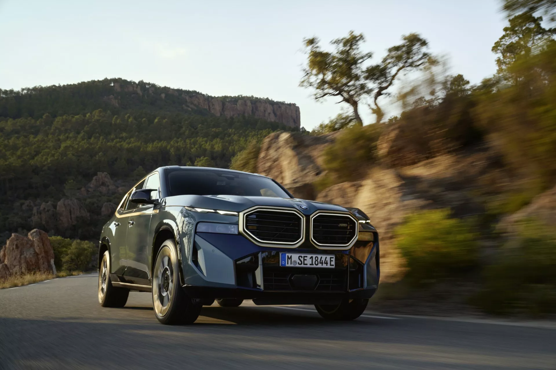 Dynamický záber luxusného vozidla v pohybe, ukazujúci jeho pokročilý aerodynamický dizajn, výraznú mriežku, štíhle LED svetlomety a športové kolesá s viacerými špicami na pozadí scenického vonkajšieho prostredia