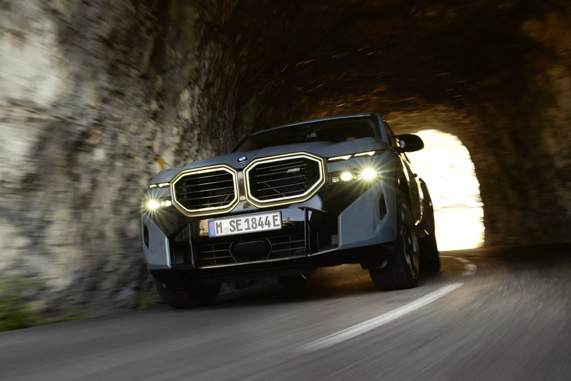 BMW XM (G09) 2023 opúšťajúce tunel, pohľad z nízkeho uhla zvýrazňujúci mriežku, LED svetlomety a dizajn kapoty v kontraste ku dennému svetlu a tieňu tunela